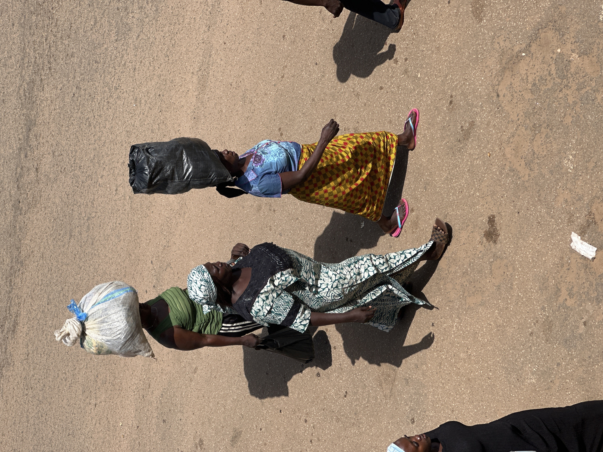 In the city of Bukuru, Nigeria, four individuals are captured in an overhead shot walking on a dusty, asphalt surface under bright daylight. A woman in the foreground, on the far left, is dressed in a black, sleeveless dress, with her left shoulder and part of her head visible. To her right, a woman walks with a patterned green and white dress and a matching headscarf. She carries a black shopping bag in her left hand and has a bundle on her head. Behind her and to the left, another woman in a green top and dark striped pants also carries a large, white, tied bundle on her head. Further to the right, a woman in a blue patterned top and a yellow and red patterned skirt walks, balancing a large black bag on her head. Her right arm is extended slightly as she walks. Part of another person's legs, wearing dark pants and sandals, is visible on the far right edge of the frame. The ground is a rough texture of asphalt with scattered small stones and a few small white paper fragments. The scene suggests a market or a busy street setting where people are carrying goods.