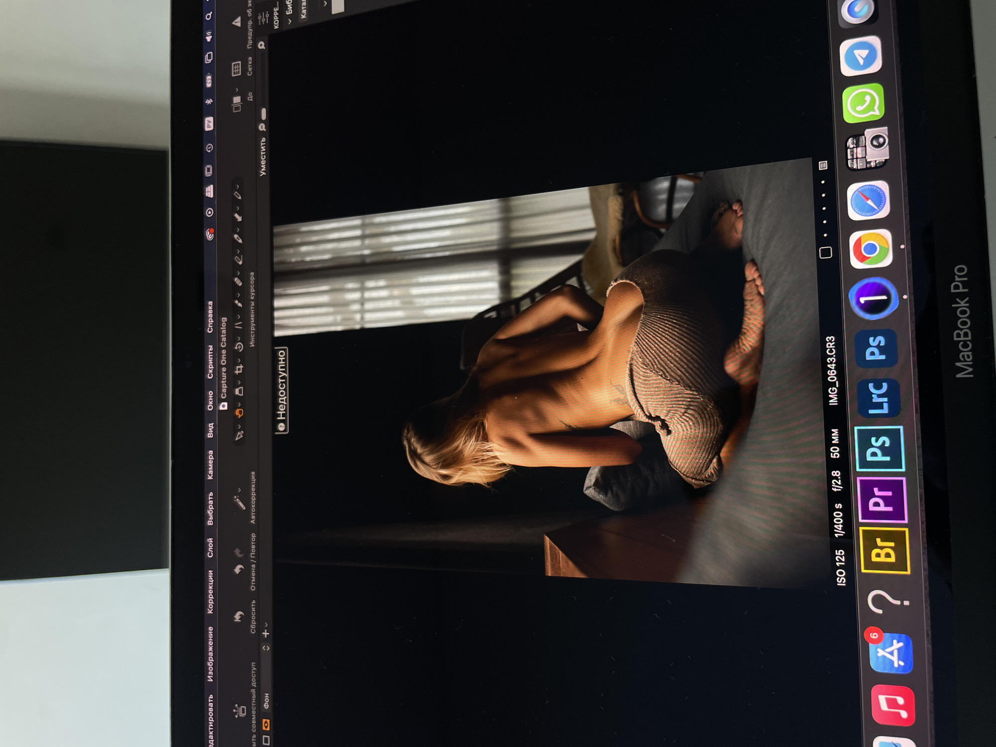 The image shows a photograph of a MacBook Pro screen, displaying a photograph of a woman lying on a bed with her back partially exposed. The operating system appears to be macOS, with various Adobe applications visible on the dock. The file name of the photo being edited is IMG_0643.CR3, shot at ISO 125, 1/400s, f/2.8, 50mm. The user interface language on the MacBook is Russian. The scene is on a desk and the city and specific location of the subject are not identifiable.