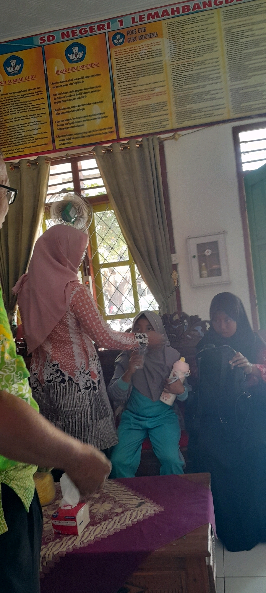 In what appears to be a classroom or community hall in Lemahbang Dewo, Indonesia, a group of individuals are gathered. A large bulletin board displays school information, including "SD NEGERI 1 LEMAHBANG," "IKRAR GURU INDONESIA," and "KODE ETIK GURU INDONESIA." Three females are the primary subjects. One adult female, wearing a pink hijab and a patterned tunic, is standing and interacting with a younger female sitting down. The younger female, also wearing a hijab and teal-colored pants, is holding a baby bottle and a cup. Another adult female, wearing a black hijab, is seated next to the younger female and appears to be holding a backpack or bag. Behind them, a patterned curtain hangs, and a fan is mounted near a window. On a wooden table in the foreground, a box of tissues is visible. The overall scene suggests a casual gathering or a moment of daily activity within the school or community setting.