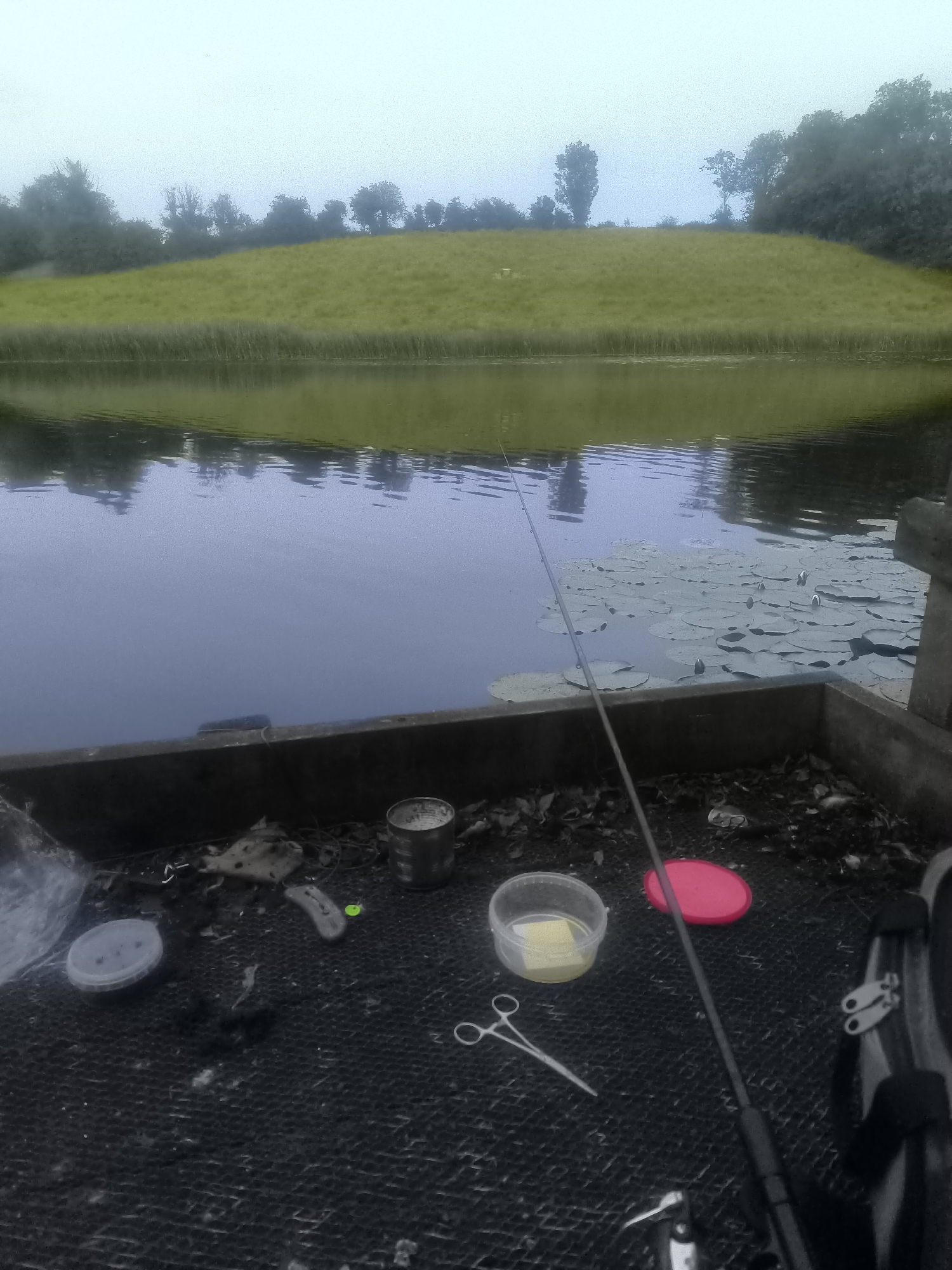 The image depicts a fishing scene at a body of water, likely a canal or river, with a grassy embankment on the far side. In the foreground, on what appears to be a wooden platform or bank, are various fishing accessories. These include a fishing rod, a small tin can, a clear plastic container with some yellow substance inside, a red lid, and a pair of scissors. The water surface is calm, with lily pads visible in the mid-ground. The overall atmosphere is overcast or hazy, suggesting it might be early morning or a cloudy day. There are no people visible in the frame. The location is identified as Roslea / Rosslea, United Kingdom.