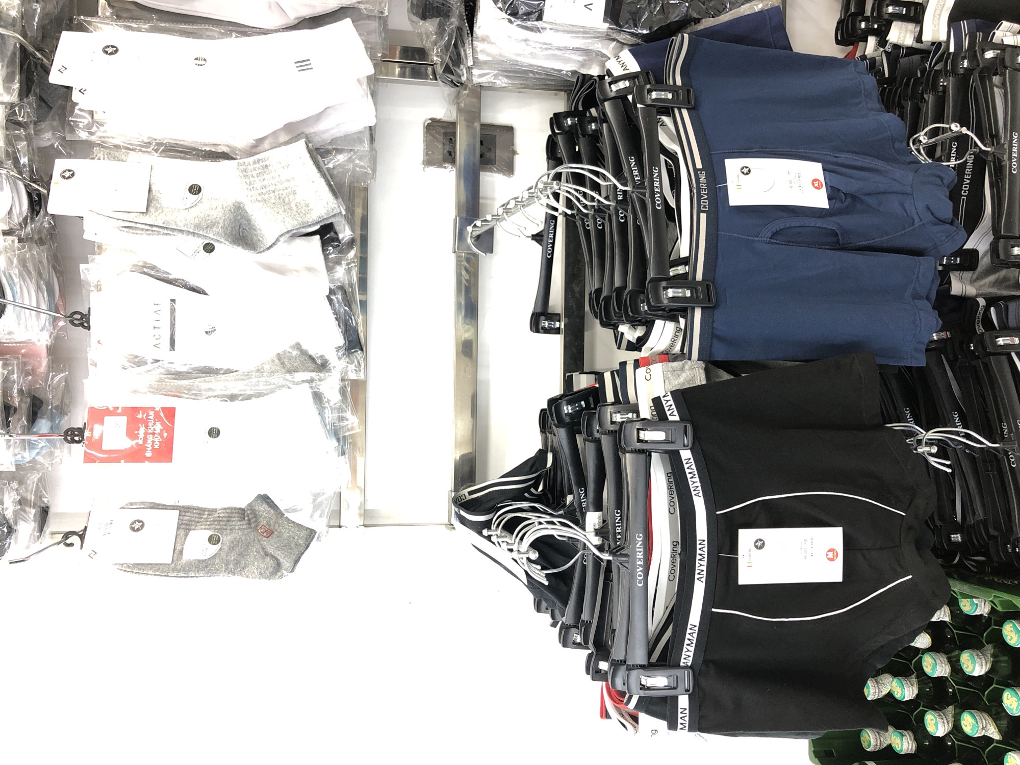 This is a vertical, eye-level shot of a clothing display in a store. The scene is well-lit, suggesting it is daytime and indoors. The display features rows of men's underwear and socks neatly arranged on hangers and in packaging.
On the left side of the image, several pairs of socks are visible, some in clear plastic packaging. These include white socks with grey accents and ribbed grey socks. Further down, a pair of white socks with the "ACTIVE" logo is displayed.
The right side of the image showcases a collection of men's boxer briefs, predominantly in dark blue and black. These are hung on black hangers, many of which are branded with text such as "COVERING" and "ANYMAN." Some of the boxer briefs have striped waistbands in white and black or white and blue. A prominent tag on a blue pair reads "COVERING" and has a red "XL" size indicator. Another tag on a black pair also shows branding and size information.
In the background, a metal shelf and a white wall with electrical outlets are visible. The lower right corner of the image shows a shelf filled with what appear to be bottles with green labels and gold caps, possibly a promotional display for beverages. The overall impression is of an organized retail environment presenting men's apparel.