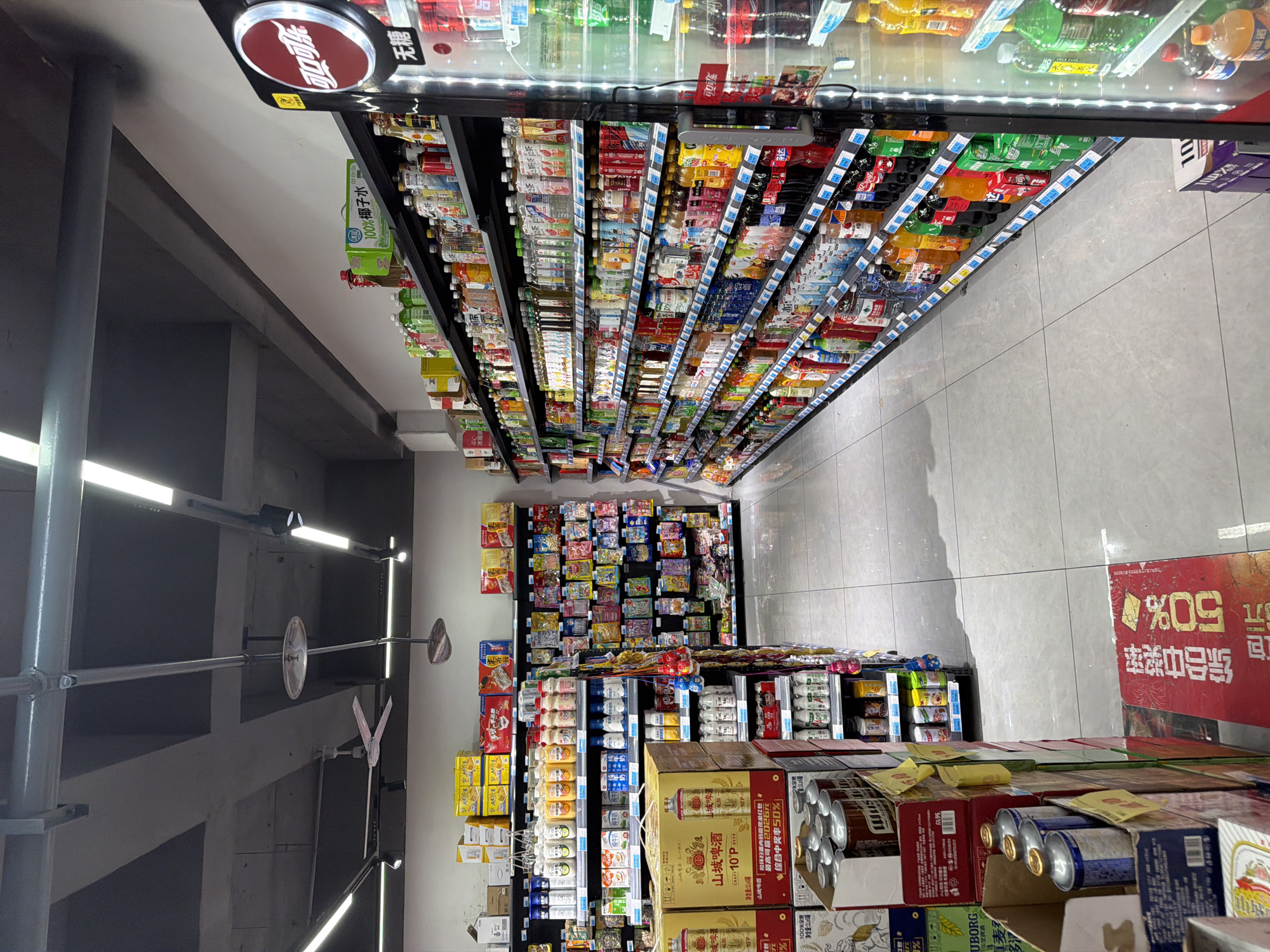 This is a medium-wide shot of a grocery store aisle in Jiangjin, China, with a focus on beverage products displayed on shelves. The aisle is well-lit with overhead fluorescent lights and illuminated refrigerators. On the left, shelves are stacked with boxes and cans of beverages, including what appear to be beer and other drinks. A large red sign on the floor in the foreground indicates a "50% off" promotion in Chinese characters. To the right, a refrigerated display case is filled with a variety of bottled and canned drinks, some with prominent branding. The shelves are organized and stocked, suggesting a typical retail environment. The flooring is tiled and clean, reflecting the lighting. The ceiling shows exposed industrial elements, including pipes and a ceiling fan, indicating a practical rather than decorative design for the store's infrastructure.