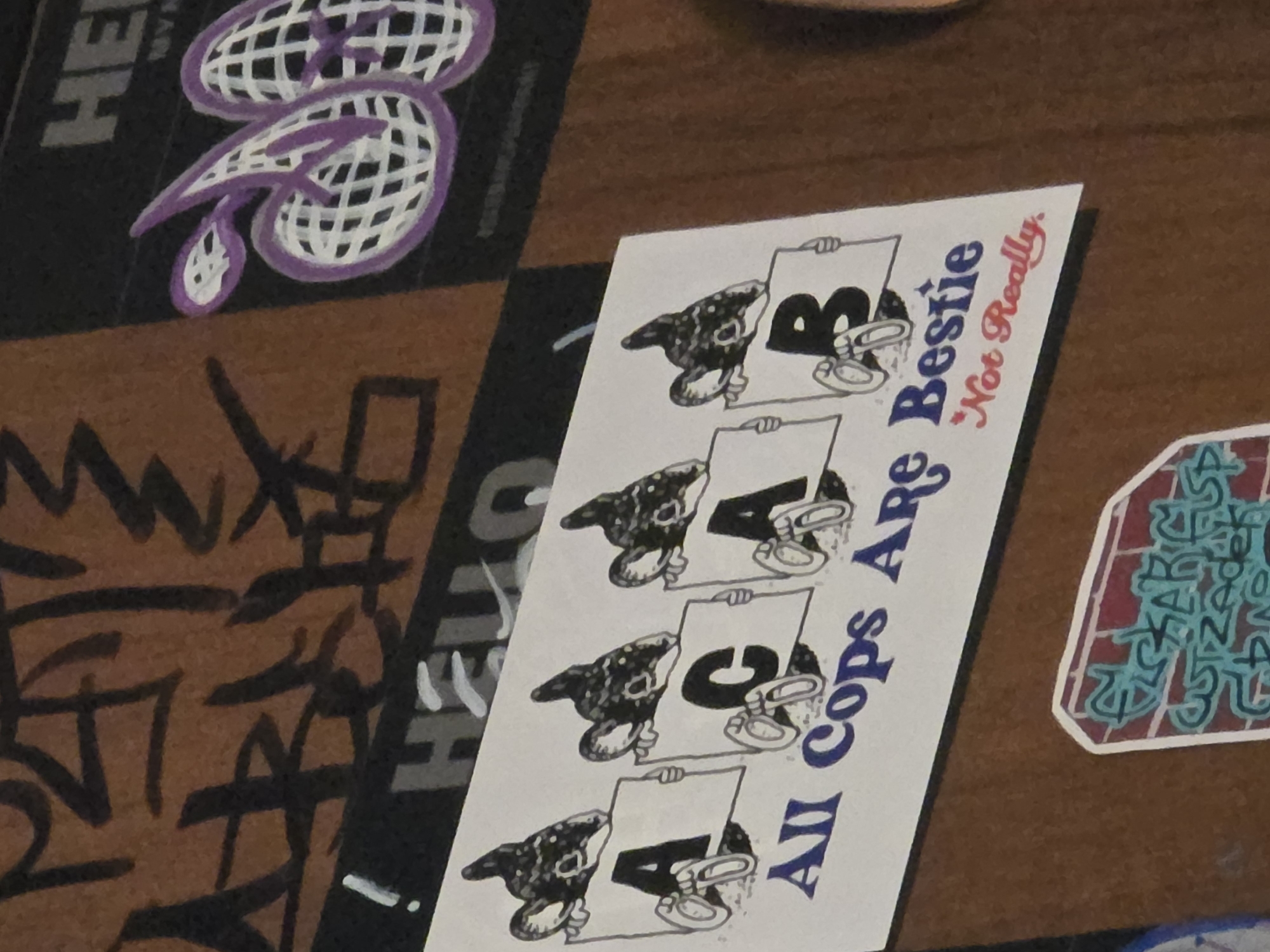 The image shows a close-up of a wall covered in graffiti and stickers. In the center is a sticker with a black and white illustration of four cartoonish mice holding signs that spell out "ACAB" and "B". Below this, the words "All Cops Are Bestie" are written in blue, followed by "Not Really" in red cursive. To the right of this central sticker, another sticker features a purple and white design of what appears to be a stylized globe or net-like pattern. The background consists of brown wood paneling with various tags and graffiti in black and white. A sticker with red and blue graffiti, appearing to depict bricks, is visible at the bottom left. The lighting suggests an indoor environment.