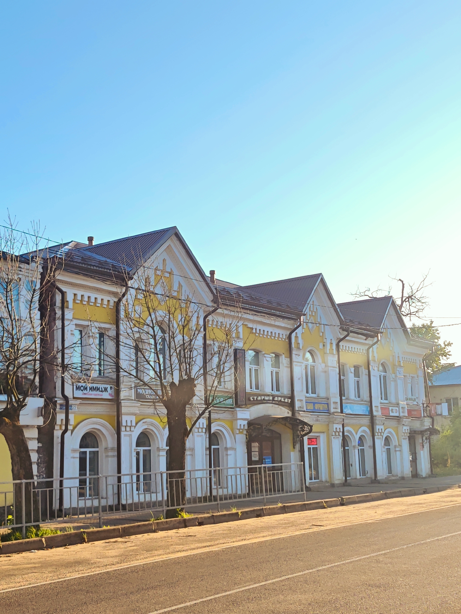 The image depicts a street view in the city of Kimry, Russia. A multi-story building with a yellow and white facade stands prominently on the right side of the frame. The building features several arched windows and ornate architectural details. A dark-colored metal roof caps the structure. Bare trees with intricate branches are situated in front of the building, suggesting a cooler season. A metal fence runs along the sidewalk, separating it from the street. The street itself is paved and appears to be a road with visible lane markings. Sunlight casts long shadows, indicating it is either early morning or late afternoon. The sky is clear and a bright blue. Several signs with Cyrillic text are visible on the building's facade, indicating shops or businesses. No people are clearly visible in the image. The overall scene is one of an urban environment with a focus on the architectural character of the building.