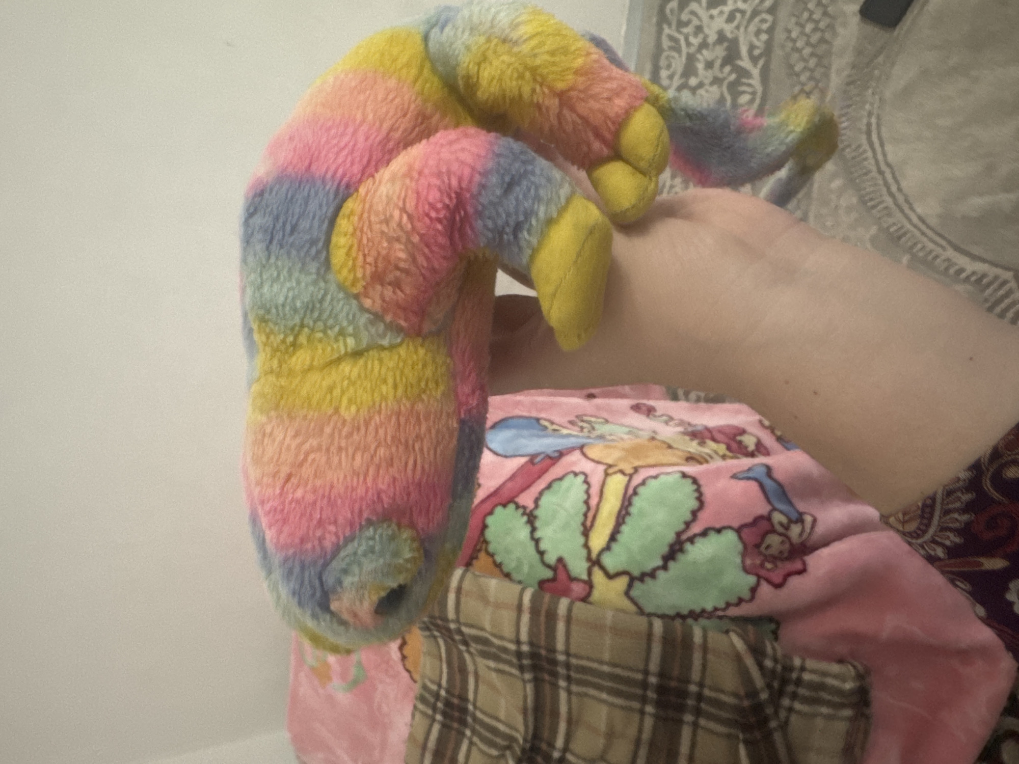 A plush toy, possibly a chameleon, with a tie-dye pattern of pink, yellow, blue, and gray, is held by a person's arm. The toy is positioned on top of blankets, one of which is pink and features cartoon characters, and another with a brown plaid pattern. The background is a plain, light-colored wall. The setting appears to be indoors, in a residential environment. No specific location in a city is identifiable.