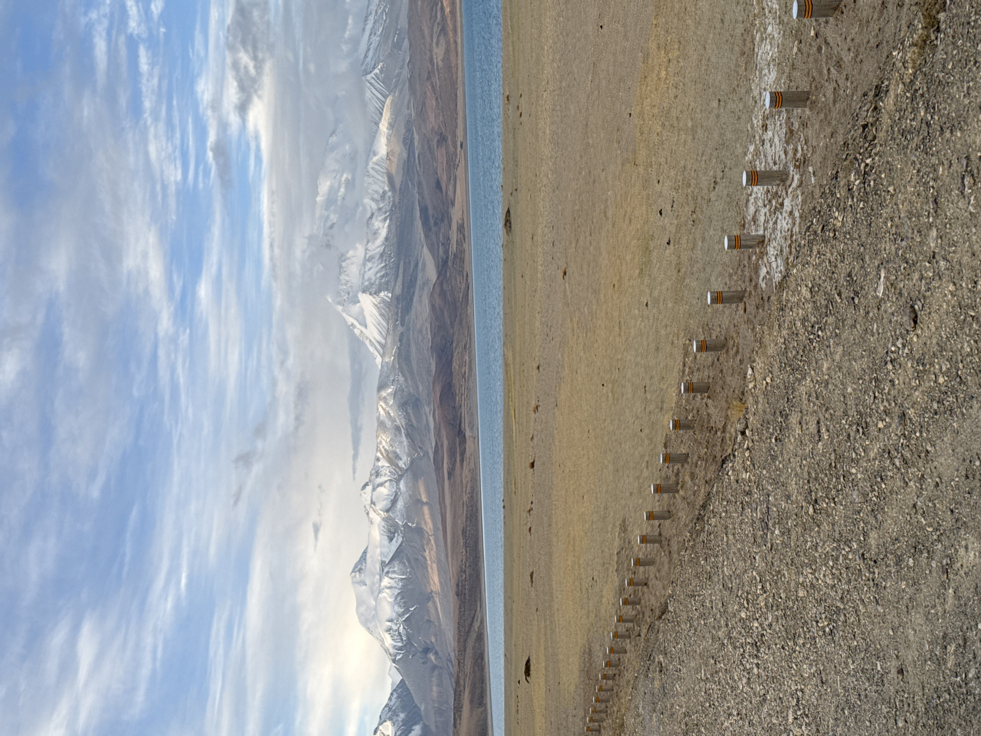 The image depicts a landscape in Purang, China, featuring snow-capped mountains, a body of water, and a shoreline marked by a series of posts. The mountains are visible on the left side of the image, with snow covering their peaks and visible cloud cover. The water body runs parallel to the mountains. The shoreline is composed of a mix of terrain. A row of evenly spaced, short, cylindrical posts runs along the shoreline and into the landscape. The posts have a distinct ringed marking. The sky above shows a gradient of blue hues interspersed with white cloud formations.