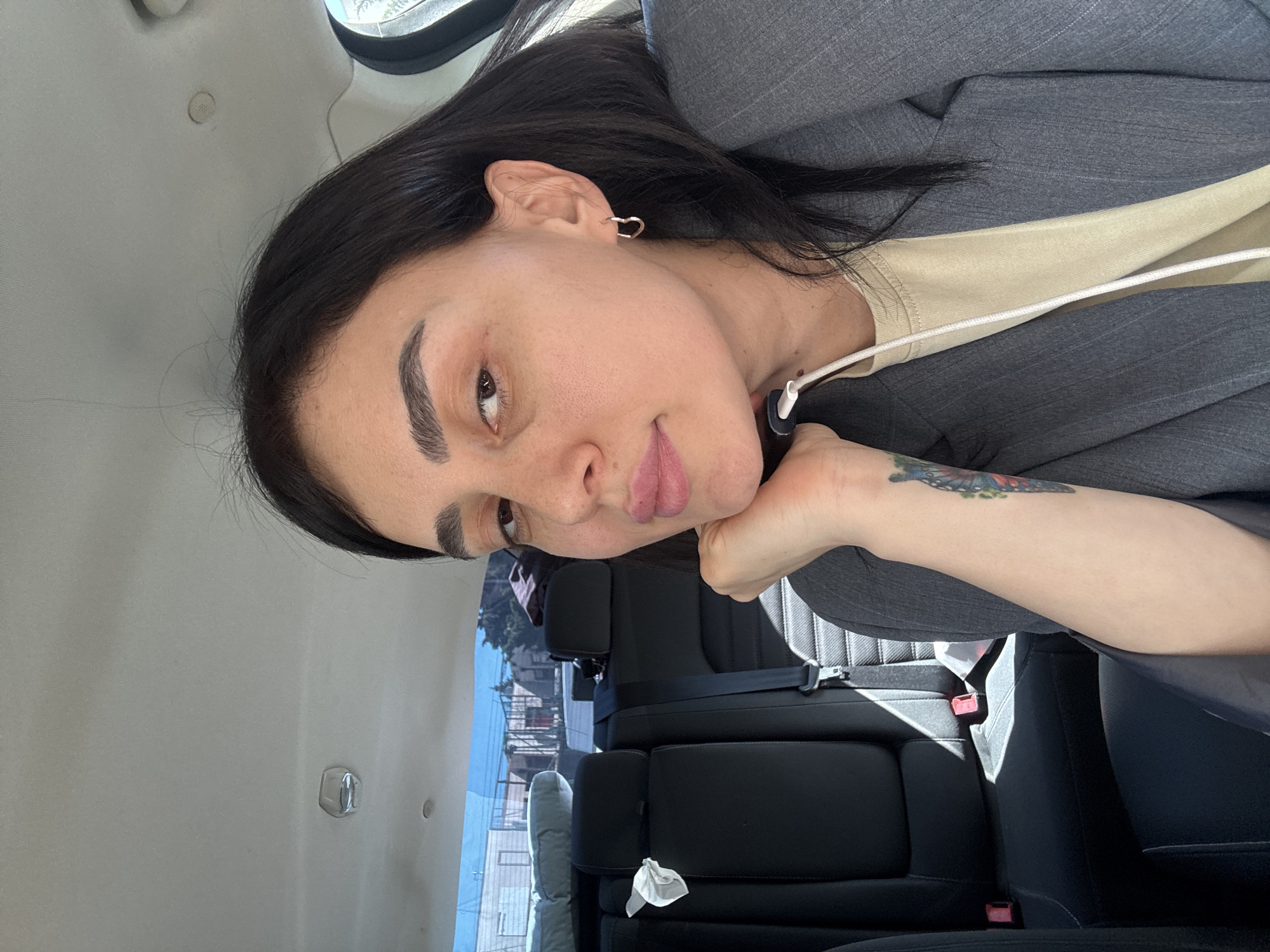 A woman with dark hair and light skin is in the back seat of a car. She is wearing a gray blazer over a light-colored shirt. Her left hand is under her chin. She has a tattoo on her left forearm. The car is stopped, and through the window, buildings can be seen. Based on the architecture visible, the location is potentially in the city of Tbilisi, Georgia. A tissue sits on the seat next to her. The car's interior is gray and black.