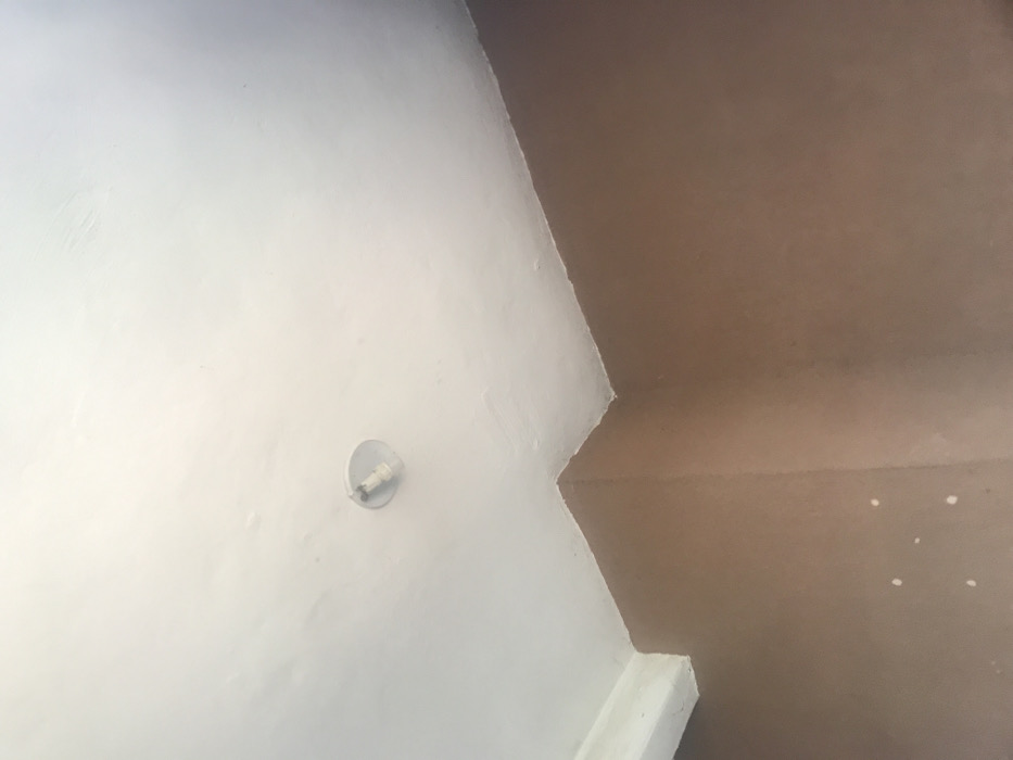 The image captures an interior architectural corner, primarily featuring two intersecting surfaces painted in contrasting colors: white and brown/tan. The dominant feature is a white wall, occupying the left and central parts of the frame. Mounted on this white wall, slightly below the center, is a simple, circular light fixture, likely a bulb holder, with a metallic component. The white wall exhibits some textural variations and minor unevenness in its paint finish.
To the right and top of the white wall, a brown or tan-colored surface extends, forming a distinct corner. This brown surface appears to be a ceiling or an upper wall segment. It also shows textural qualities and contains a few small, scattered white specks, possibly paint splatters or debris, particularly in the lower right portion of its area.
In the bottom right corner of the image, an additional white, rectangular structural element or trim is visible, indicating another architectural junction. The lighting is even, suggesting an indoor setting. There are no people or discernible activities taking place, and no visible text within the frame. The architectural elements presented are generic and do not offer specific visual cues that uniquely identify the stated location of Umudike, Nigeria.