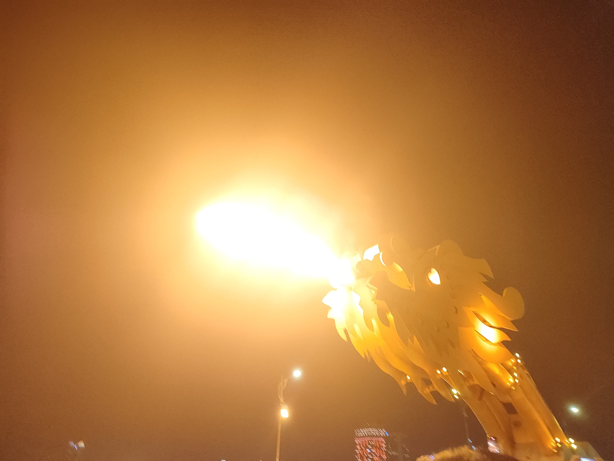 The image depicts the head of the Đà Nẵng Dragon Bridge in Vietnam, breathing fire. The fire is intensely bright, obscuring details of the dragon's head, though its overall shape is discernible. The dragon's head is adorned with decorative elements and small lights. The surrounding environment is dark, suggesting a nighttime scene. Streetlights and building lights are visible in the background, indicating an urban setting. A crowd of people is visible at the base of the bridge.