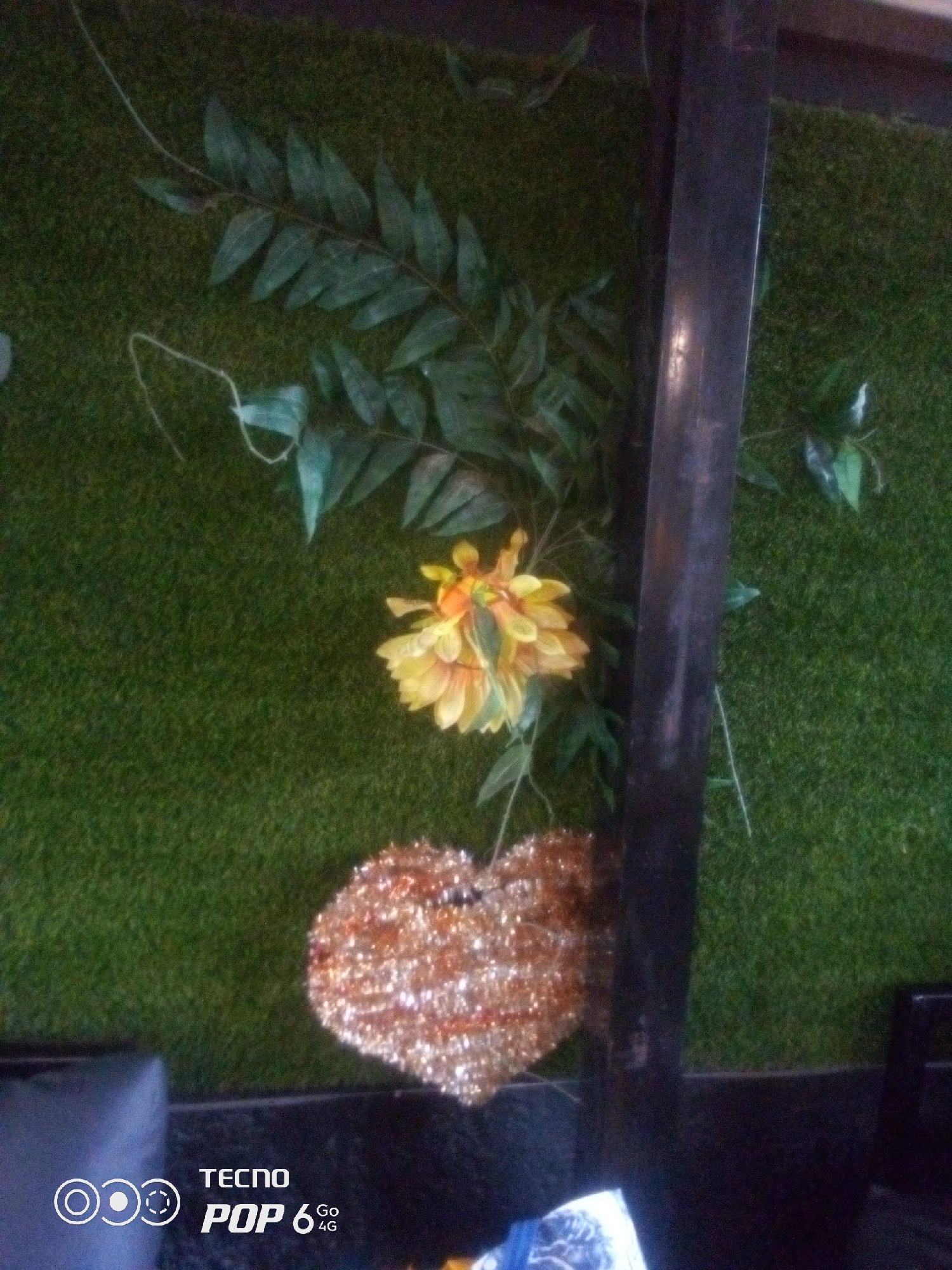 The image shows a vertical metal pole against a green, textured background that resembles artificial grass. A single artificial yellow flower with orange accents is positioned to the left of the pole, amidst artificial green leaves and tendrils. Below the flower and to the left of the pole, a heart-shaped object covered in gold and copper sequins is visible. The bottom left corner of the image contains a watermark with "TECNO POP 6 Go 4G" and a circular design. The scene appears to be indoors, possibly in a commercial establishment or a decorated space in Howayi, Nigeria.