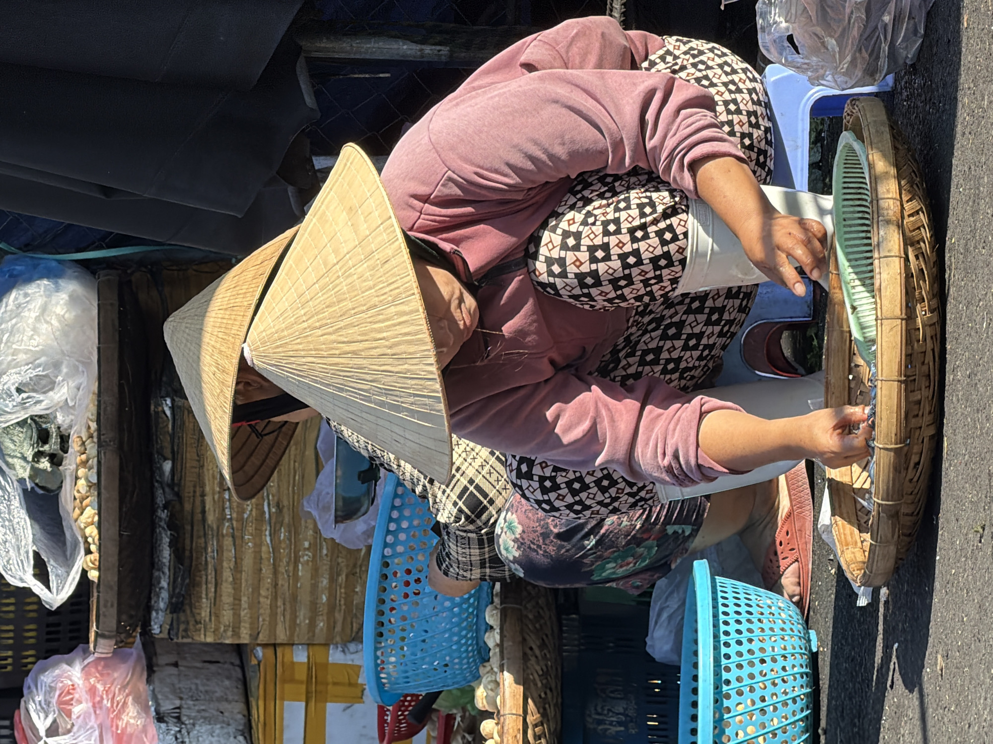The image captures a street scene in Nha Trang, Vietnam, likely a market. Two women, both wearing conical Asian hats, are present. The woman in the foreground is crouched, wearing a pink sweatshirt and a patterned skirt. Her hands are engaged with a woven basket in front of her, which appears to contain small fish or seafood. A light green plastic colander or sieve is also in the basket. White boots are on her feet. Beside her is a blue plastic basket filled with what looks like produce, possibly shallots or small onions. Behind her, another woman in a similar hat is partially visible, wearing a checkered top and a patterned skirt, and is also crouched. The background shows stacks of goods, including what seems to be crates and a large black fabric, suggesting a commercial area. The ground is paved with asphalt, and sunlight casts distinct shadows, indicating an outdoor setting.