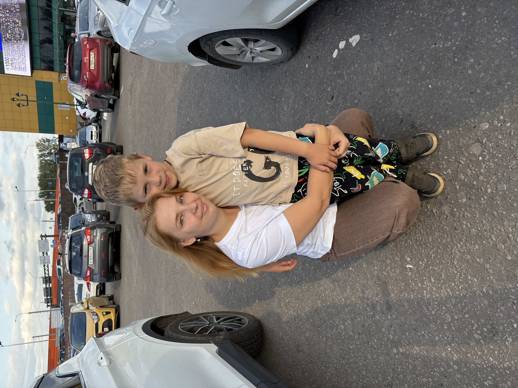 A mother and her young son are posing for a photo in a parking lot. The mother is lying on her back with her head propped up on her arm, smiling at the camera. Her son is lying on top of her, also looking at the camera and smiling. He has his arms wrapped around her neck. Both are casually dressed. The mother is wearing a white t-shirt and brown pants, and the son is wearing a tan t-shirt with a graphic print and patterned pants. They are in a parking lot with several cars visible in the background. Some of the cars have visible license plates, with text including "E789CH 76" and "M169cc 76". A large building with a banner displaying "VTB.RU" and "1000" is visible behind the cars. The sky is overcast, suggesting it might be daytime with cloudy weather. The scene appears to be a casual outing, possibly after shopping or running errands.