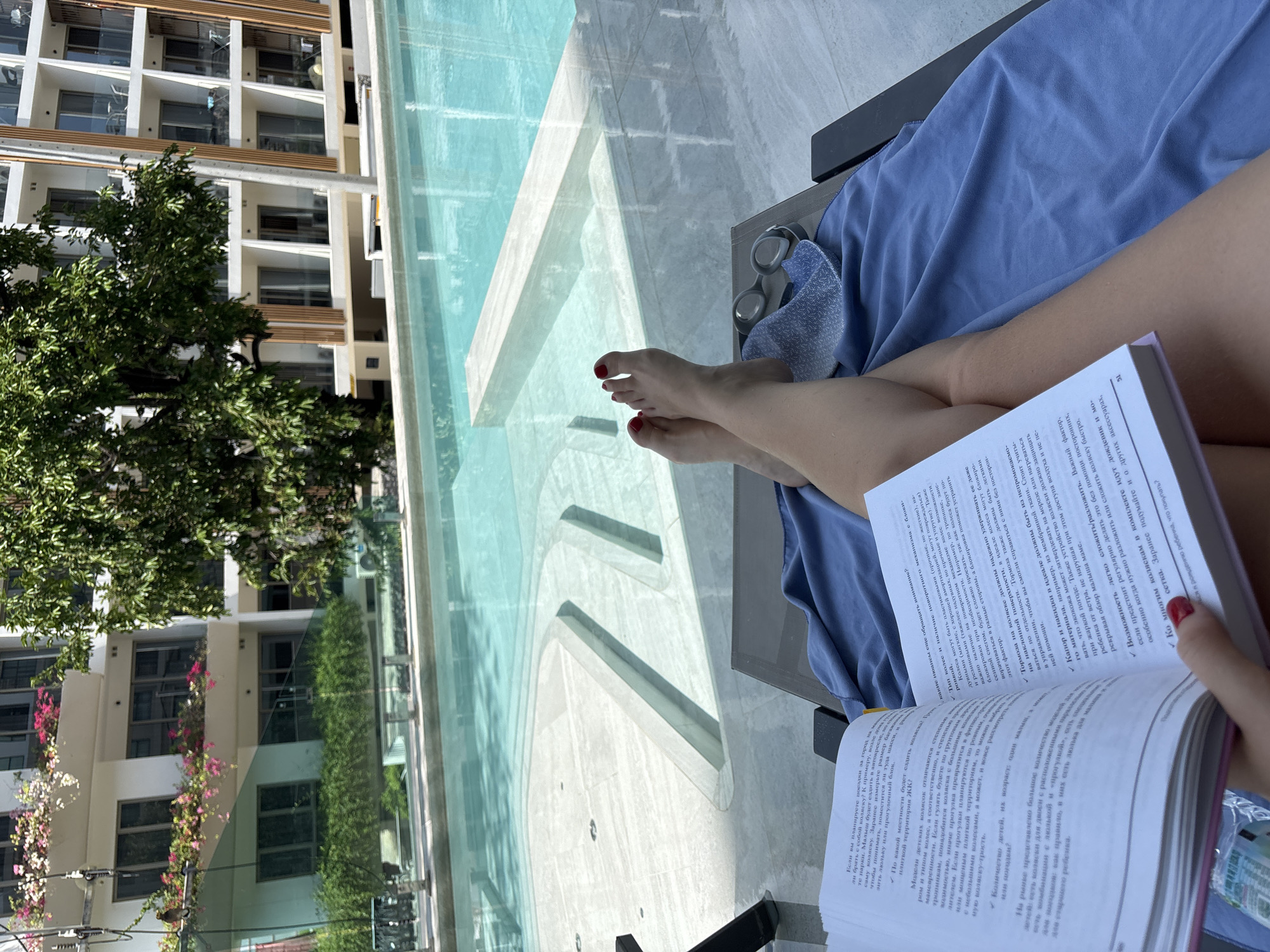The image captures a first-person perspective of a person relaxing by an infinity pool, reading a book in bright daylight. The scene suggests a leisurely vacation or resort setting.
The main subject is a person, whose legs, feet, and one arm are visible. The person is lying on a dark lounge chair, covered partially by a light blue towel or sarong. Their toenails and fingernails are painted red. A pair of swimming goggles rests on the lounge chair next to them. The person is holding an open book with white pages and black text, actively reading.
The background features a clear blue infinity swimming pool with light-colored tiles, reflecting the sunny sky. Beyond the pool, several modern, multi-story buildings with balconies are visible, likely part of a hotel or apartment complex. Lush green trees and some flowering plants (pink and white) add to the tropical ambiance. The overall impression is one of relaxation and calm.
The visible text in the open book is in Russian. It appears to be a guide or advice related to choosing baby strollers, discussing features like wheel types ("Тип колес"), braking systems, weather protection ("Капор и накидки"), and ease of folding ("Возможность легко сложить/разложить"). One heading visible is "По какой местности будет ездить малыш?", and the page number 51 is clearly seen at the top right.
Given the location context of Choeng Thale, Thailand, the scene perfectly depicts a warm, sunny day at a resort or hotel in a tropical environment.