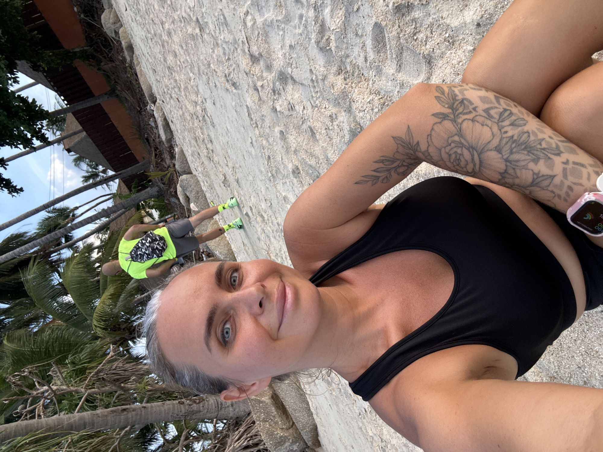 A woman with blonde hair pulled back in a bun and blue eyes smiles at the camera. She is wearing a black sports bra and black shorts. She has a tattoo of flowers and leaves on her left arm. In the background, a man with a shaved head is walking away from the camera. He is wearing a neon yellow t-shirt and gray shorts, with a black and white patterned backpack. He is also wearing neon green athletic shoes. They are on a sandy beach with palm trees and some rocks. In the distance, there are buildings with thatched roofs. The sky is partly cloudy. The scene appears to be in the Ko Samui District of Thailand, given the tropical vegetation and architectural style.