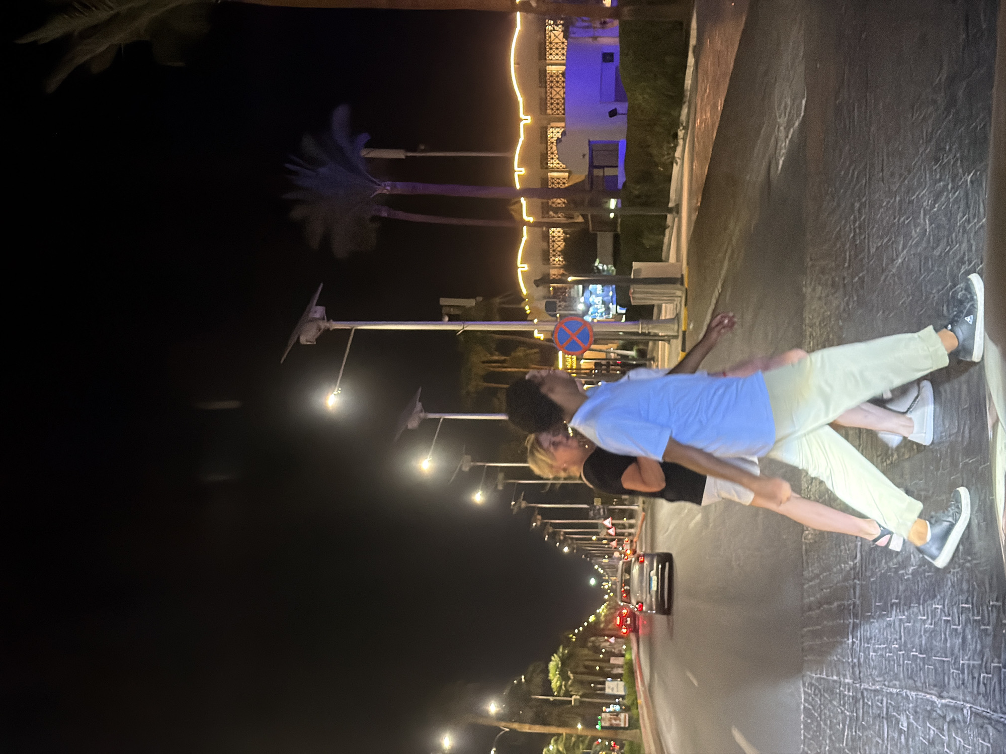 The image depicts a nighttime scene on a street in Sharm El Sheikh, Egypt. Two people are crossing a road at a pedestrian crossing. The person in the foreground, appearing to be of male gender, wears a light blue short-sleeved shirt and light-colored pants, and dark sneakers. They are walking from left to right across the frame. Behind them, a person who appears to be female, wearing a black t-shirt and light shorts, is also crossing, walking in the same direction. Several cars are visible on the road, with headlights and taillights illuminated. The street is lined with palm trees and lampposts, some of which are solar-powered. Buildings with illuminated decorative lighting are visible on the right side of the street. The sky is dark, indicating it is nighttime. The ground of the pedestrian crossing appears to be textured.