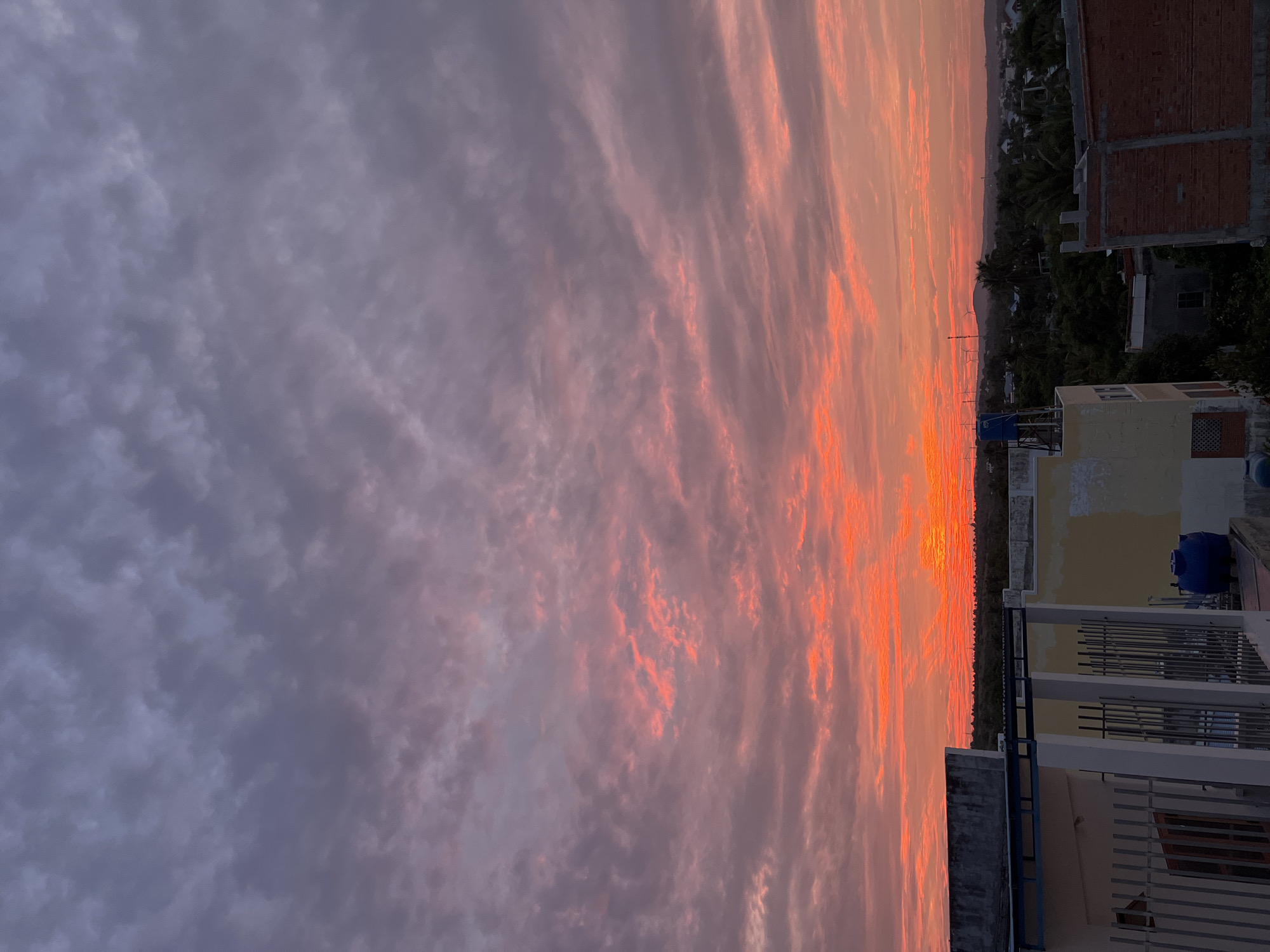 This image captures a vibrant sunset over the city of Mũi Né, Vietnam. The sky is a dramatic canvas of color, with fiery oranges and reds near the horizon blending into soft pinks and purples higher up. Wispy clouds are illuminated by the setting sun, creating a textured and dynamic ceiling. In the foreground, the rooftops of several buildings are visible. One building, partially in shadow, features a light-colored facade and metal fencing. Adjacent to it, another building displays a yellow painted exterior, and a blue water tank is perched on its roof. Further into the scene, a red brick structure is under construction. Beyond the buildings, a dense canopy of green trees stretches across the landscape, interspersed with glimpses of smaller houses. In the distance, the faint outlines of hills and what appear to be wind turbines can be seen against the colorful sky. The overall impression is one of a peaceful, rural or semi-urban setting at dusk.