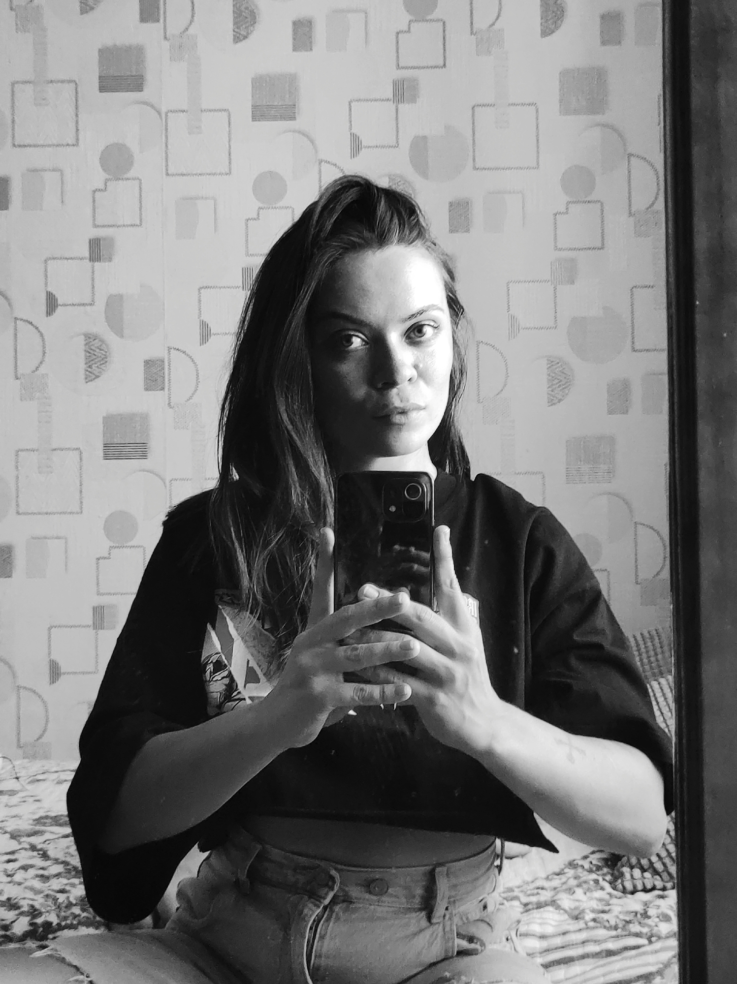 A black and white portrait shows a young woman in what appears to be a bedroom. She is holding up a smartphone, taking a selfie in front of a mirror. Her dark hair is partially obscuring her forehead. She is wearing a dark, short-sleeved shirt that reveals part of her midriff, and light-colored shorts. Behind her, the wallpaper features a geometric pattern of squares, circles, and lines. A bed with a patterned bedspread is visible below her.
Given the information in the prompt, this scene is assumed to be in Chaikovsky, Russia, although there is no concrete evidence of this in the image itself.