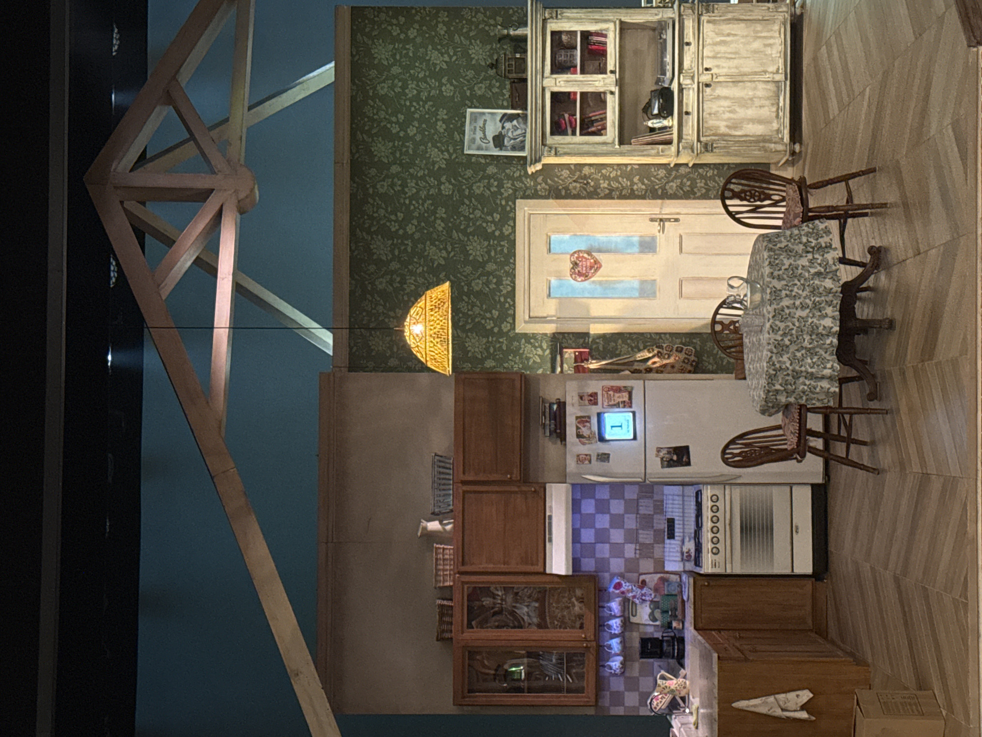 This media file displays a detailed, vertically oriented image of what appears to be a miniature set or a stage prop depicting a cozy domestic interior, possibly a kitchen and dining area. The image is rotated 90 degrees clockwise from its natural orientation.
The scene features an inviting home environment, rich in detail. To the left (when correctly oriented), a large, intricate wooden truss structure with exposed beams is visible against a dark teal background, suggesting a loft, attic, or stage rigging.
The main interior space is floored with light wood planks laid diagonally in the dining area, transitioning to a checkered tiled floor in the kitchen zone. The walls are covered in dark green wallpaper with a subtle floral pattern.
In the dining area, a round wooden table is draped with a floral tablecloth, accompanied by two classic wooden spindle-back chairs. Above the table, a unique, ornate pendant light fixture with a warm glow illuminates the space. Adjacent to the table, an antique-style white wooden hutch with glass-paned upper doors filled with books and various items stands against the wall. A framed picture depicting a couple, possibly from an old advertisement, is visible on the wallpaper, with the word "Cadbury's" faintly discernible. Below the hutch, a white wooden door with two glass panels features a small heart-shaped decoration in its center.
The kitchen area includes a white refrigerator adorned with magnets and small pictures, displaying the number "1" on its digital panel. A white stove with multiple burners and controls is next to it. Wooden cabinets, some with glass doors showcasing dishes, line the walls above and below a countertop area. Various kitchen items like mugs and possibly a kettle are arranged on the counter. A large brown box is situated on the floor near the kitchen cabinetry.
The overall atmosphere is one of warmth, nostalgia, and domestic comfort. No people are present, and no specific activity is taking place, but the scene implies a lived-in space. The lighting suggests an evening or indoor setting.