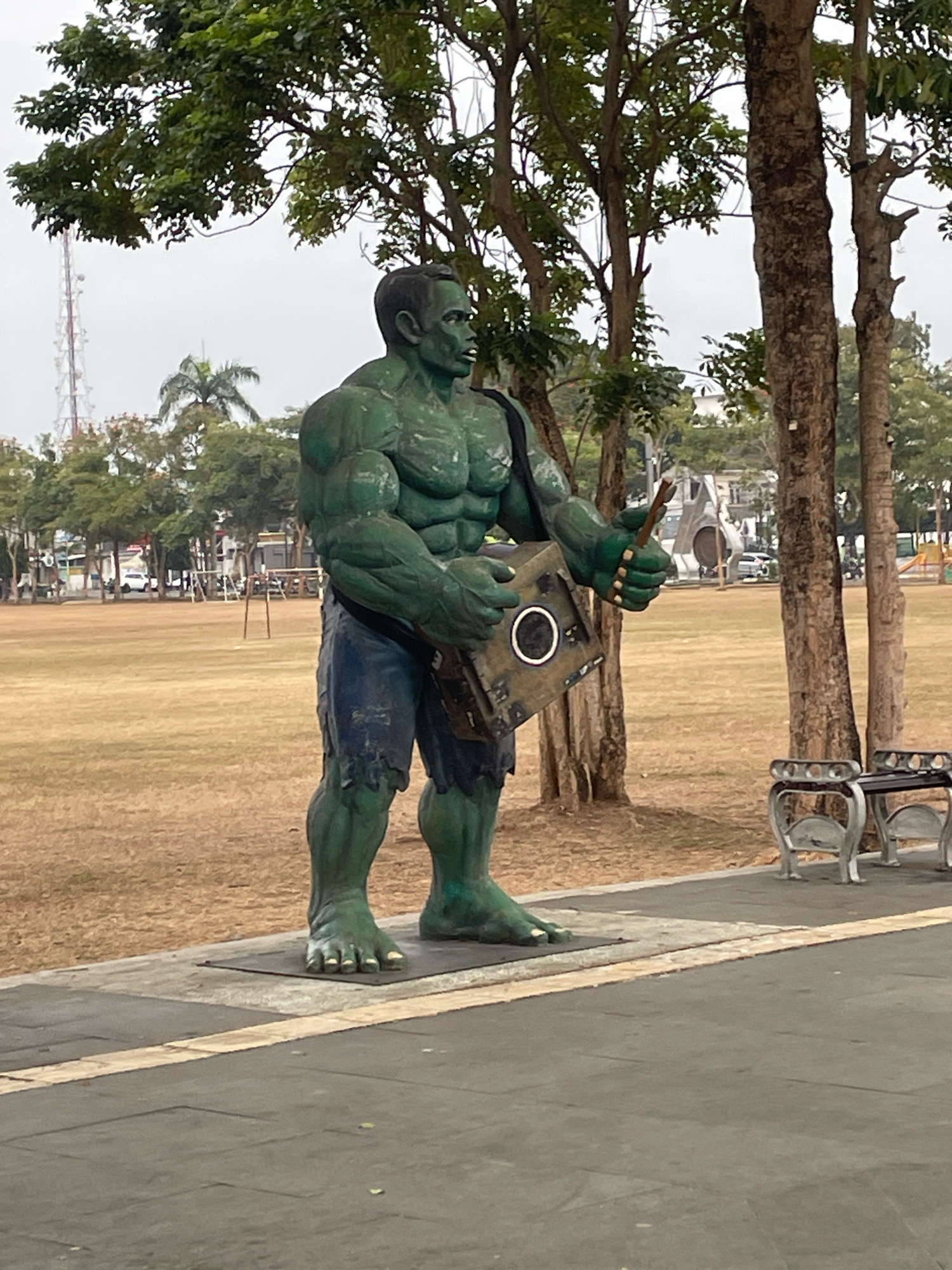 The image shows a large, green statue of the Hulk standing in an outdoor area. The statue is positioned on a paved platform, with a grassy field behind it. Trees are visible in the background, along with some buildings and a radio tower. There are two benches to the right of the statue. The Hulk statue is depicted in a muscular pose, wearing torn blue shorts. It holds a brown object in its right hand and a square, metallic object in its left arm. The scene appears to be in a public park or recreational area in Purworejo, Indonesia, given the context of the statue and its setting. The sky is overcast, suggesting a cloudy day.