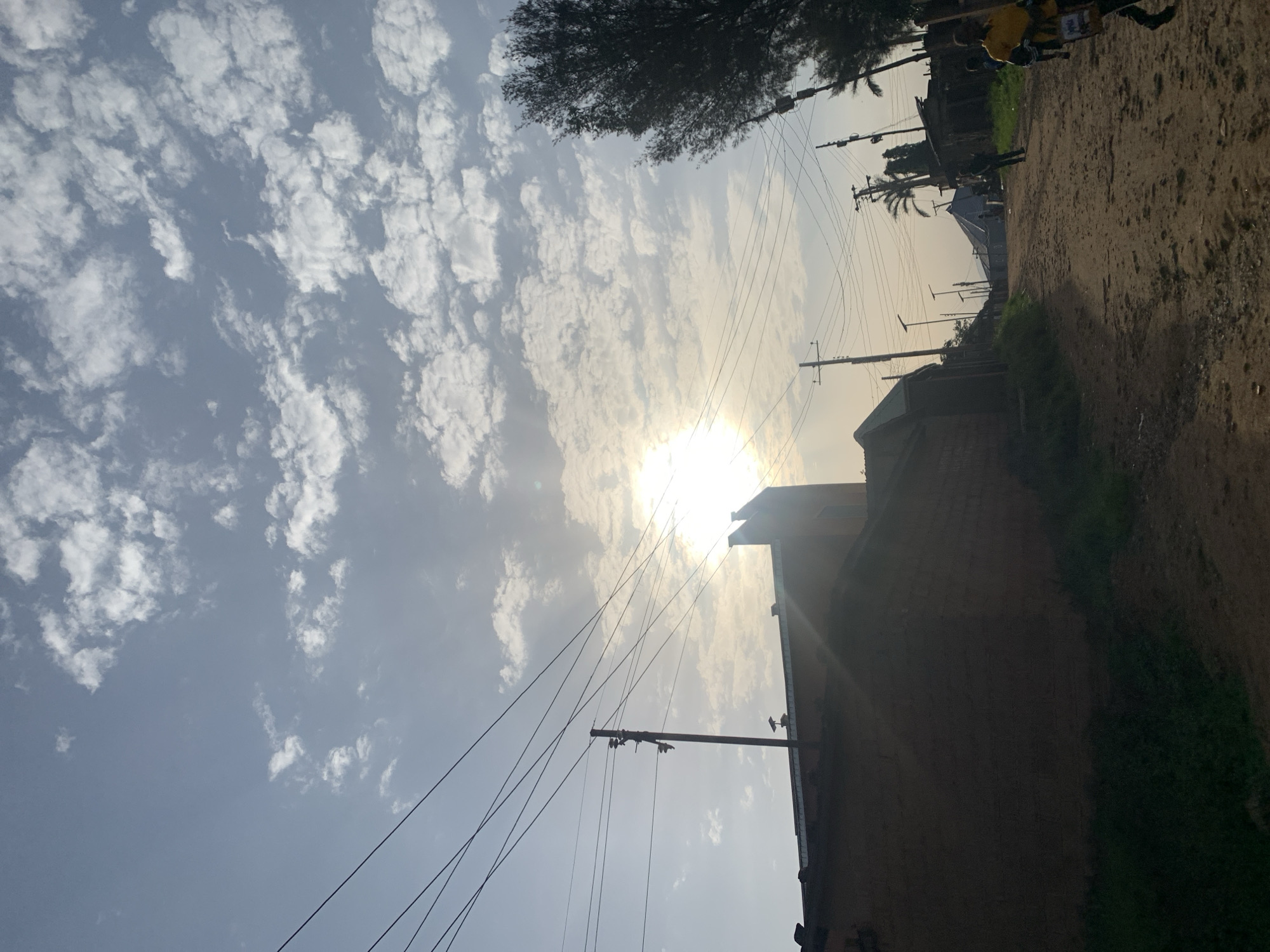 The image depicts a street scene in the city of Jos, Nigeria, characterized by a dirt road and buildings constructed with reddish-brown bricks. The sky is filled with scattered white clouds, and the sun is partially obscured, casting rays of light across the scene. Overhead, numerous electrical wires are strung between utility poles, indicating the presence of infrastructure. On the right side of the road, a tall tree with dark foliage is visible, and further down the street, several people are walking. One individual in the foreground on the right is wearing a yellow shirt and appears to be carrying a bag. The overall impression is that of a typical urban street in Jos, likely during the daytime, with a blend of residential structures, utility services, and pedestrian activity.