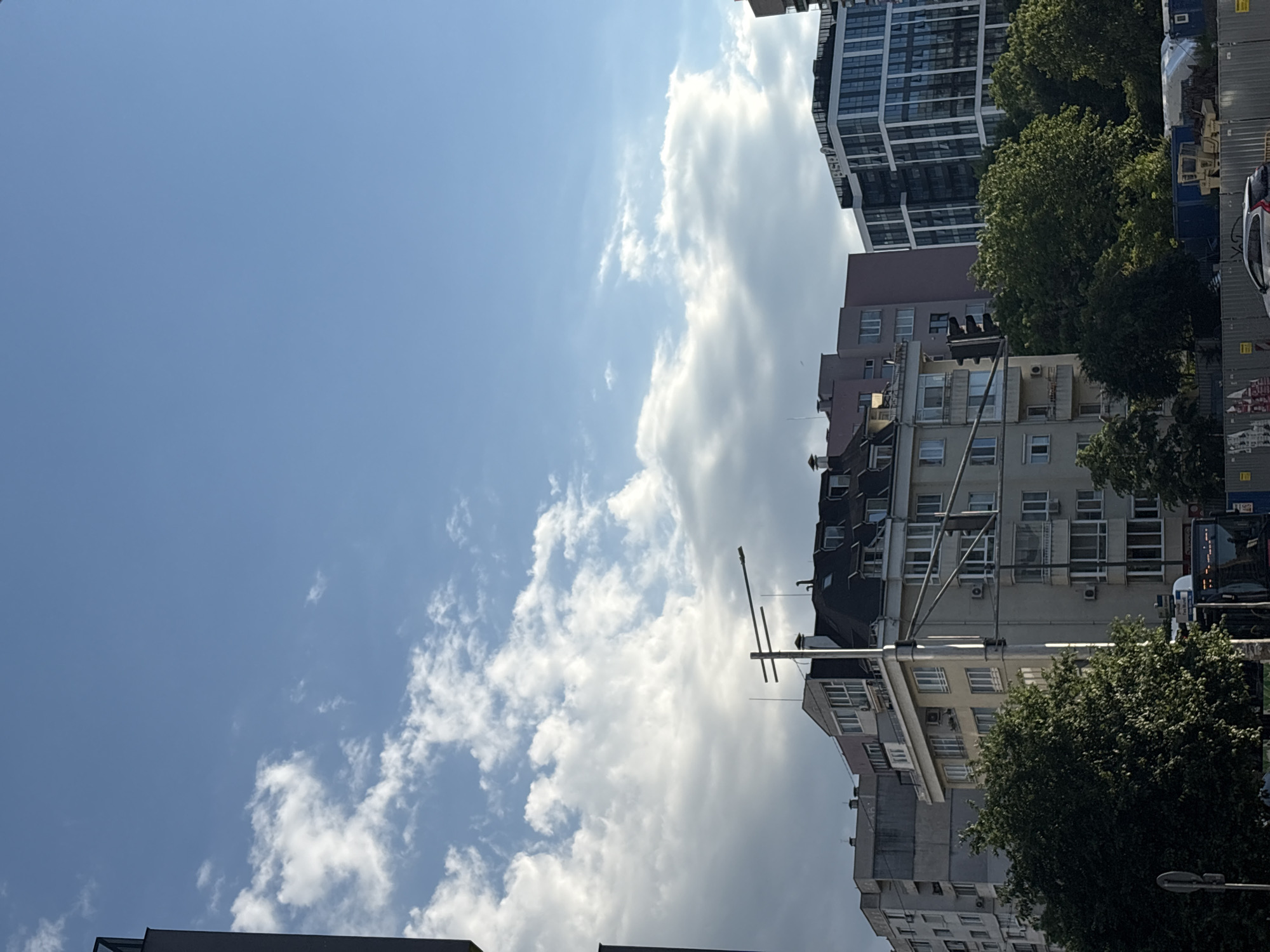 The image captures a view in the city of Sofia, Bulgaria. The scene is dominated by the sky, partially covered with white clouds. Several buildings of varying heights and architectural styles are visible, including residential blocks and a taller, modern apartment building. Trees with green foliage are present along the street. A bus is visible on the street, indicating urban traffic. Overhead cables stretch across the scene, characteristic of the urban infrastructure. The overall setting is typical of a sunny day in a European city.