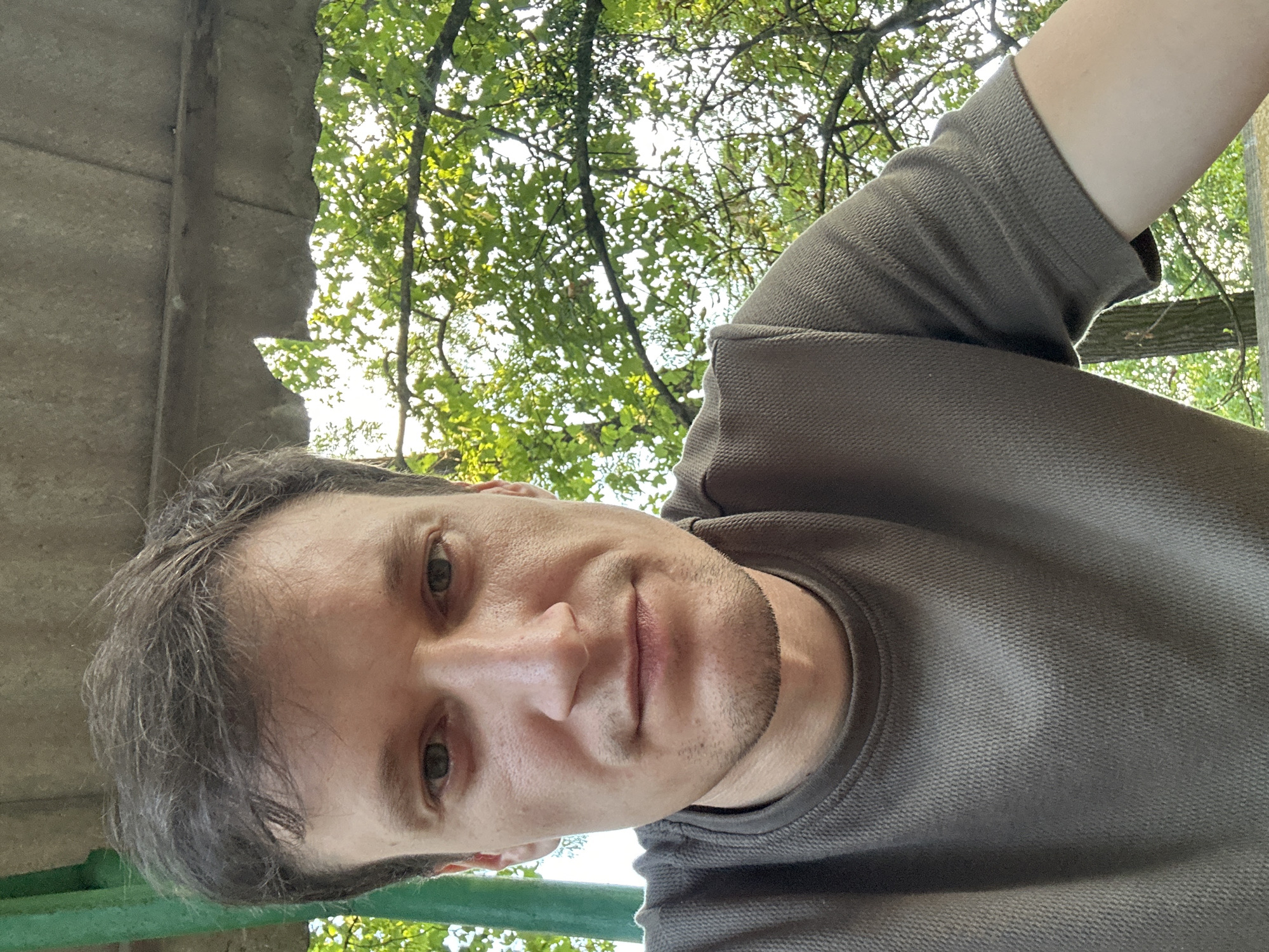 A close-up, selfie-style photograph shows a man with short, dark brown hair and light skin. He is wearing a dark olive green t-shirt with a textured weave. His head is tilted slightly down and to the left, and he is looking directly at the camera with a neutral expression. His eyes are visible, and he has a small amount of stubble on his chin and jawline.
The background is outdoors, with dappled sunlight filtering through green leaves of trees, suggesting a daytime scene. Part of a concrete structure, possibly a roof or overhang, is visible at the top of the frame. A green metal pole is present on the left side of the image, partially obscuring the man's shoulder.
The image does not provide specific visual cues to pinpoint its exact location within Makiv, Ukraine, beyond being an outdoor setting with vegetation and some form of man-made structure.