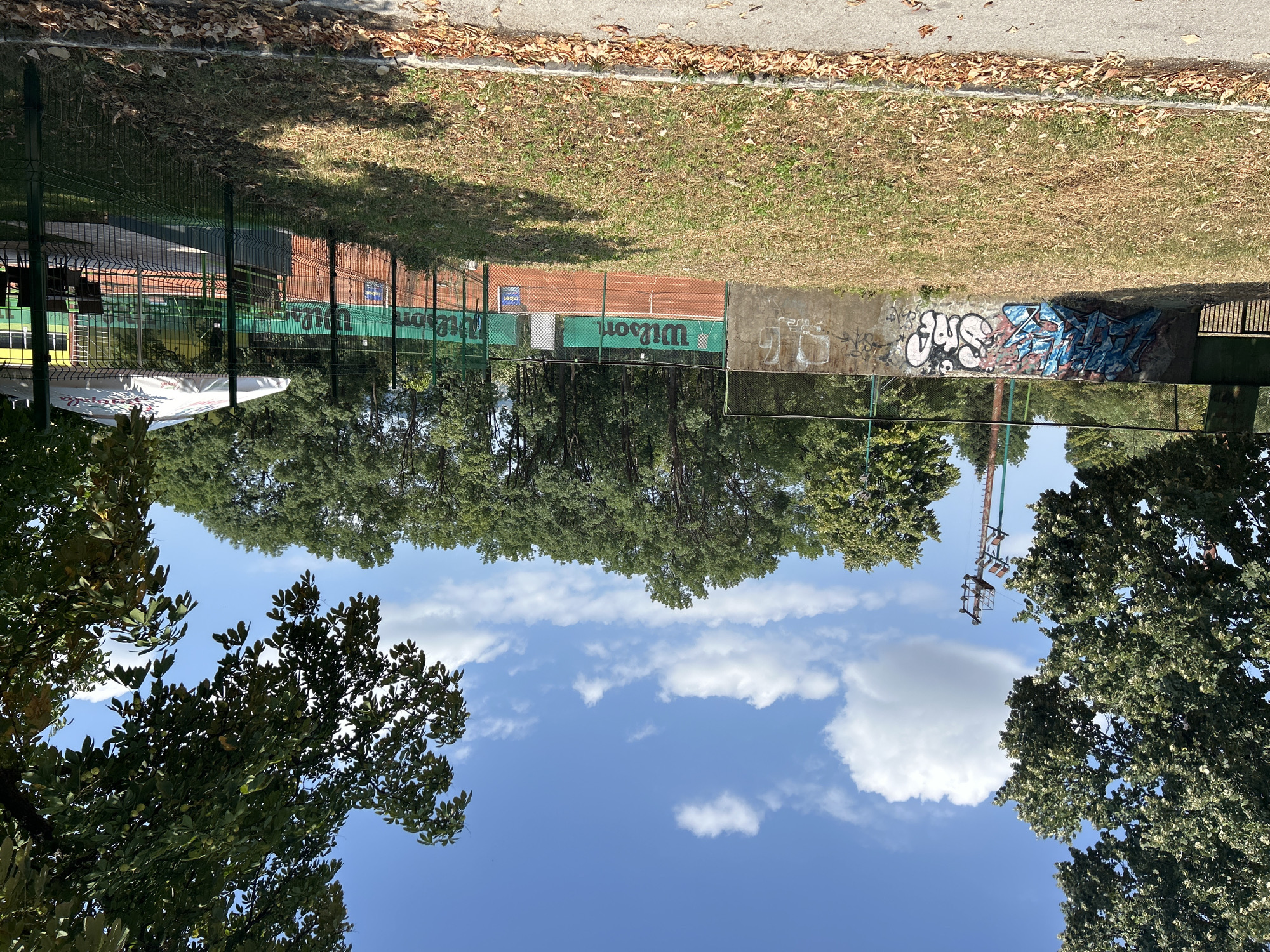 This is an outdoor, eye-level, medium shot of a tennis court on a clear day. The court has clay surfaces and green nets with "Wilson" branding. In the foreground, a dry, grassy area with scattered autumn leaves leads to a concrete wall adorned with graffiti. Beyond the tennis court, a line of trees forms a dense green backdrop against a bright blue sky with scattered white clouds. A small building with a yellow facade and a white umbrella is visible on the right side of the frame, behind a metal fence. A solitary power pole stands to the left of the tennis court, partially obscured by trees. The scene appears to be located in the city of Vratsa, Bulgaria. There are no people present in the image.