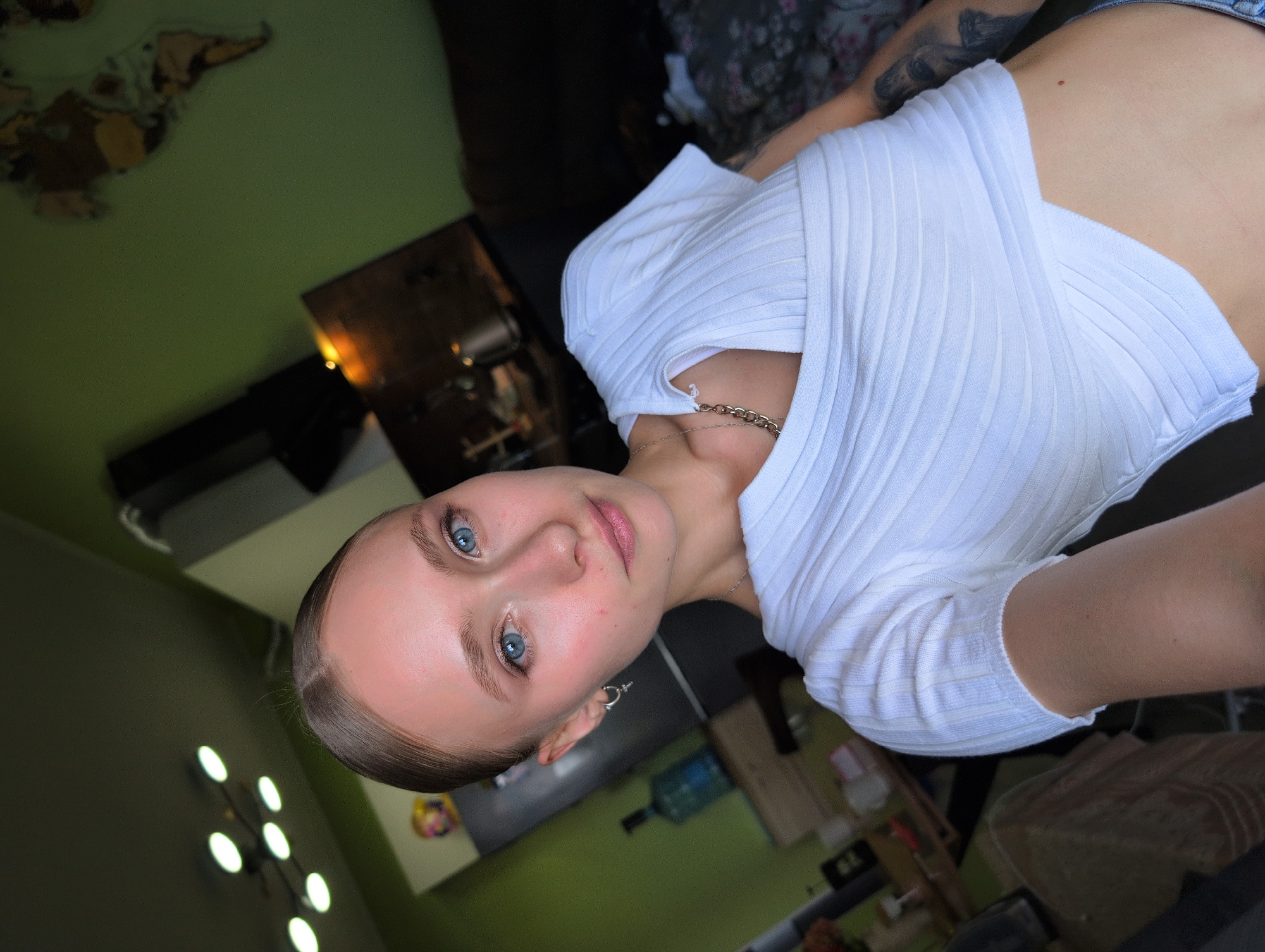 A young woman with blue eyes and blonde hair pulled back is taking a selfie in an indoor setting. She is wearing a white, ribbed, cropped top and blue jeans. A silver necklace is visible. In the background, a kitchen area with cabinets and appliances can be seen, along with a wall decorated with a world map. There is a bottle of water on a counter, and various objects on shelves. The room appears to be dimly lit, with a multi-bulb ceiling light fixture visible. The setting is consistent with an apartment in Bugry, Russia.