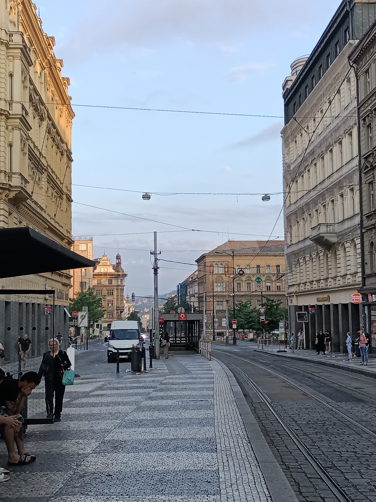The image depicts a street scene in Prague, Czechia, likely during the late afternoon or early evening. The architecture suggests an older European city with ornate buildings on either side of the street. The street is paved with cobblestones and features tram tracks running down the center.
On the left side of the street, a light-colored building with classical architectural details is visible, featuring multiple stories with arched windows and balconies. Part of a dark awning or canopy extends from the building, covering a sidewalk area. Several people are present on this sidewalk, some walking and others sitting on a bench. One person is holding a green bag with a smiley face on it.
On the right side of the street, a darker-colored building with a more modern facade on its lower floors stands. A Burger King restaurant is clearly visible on this side. People are also present on the sidewalk here, some queuing near a bus stop shelter.
In the middle ground, a white van with "IVECO" visible on its front is driving down the street. Further down the street, traffic lights are visible, and more buildings recede into the distance, with a hilly landscape in the background. The sky is overcast with some lighter patches. The overall impression is of a busy urban environment.