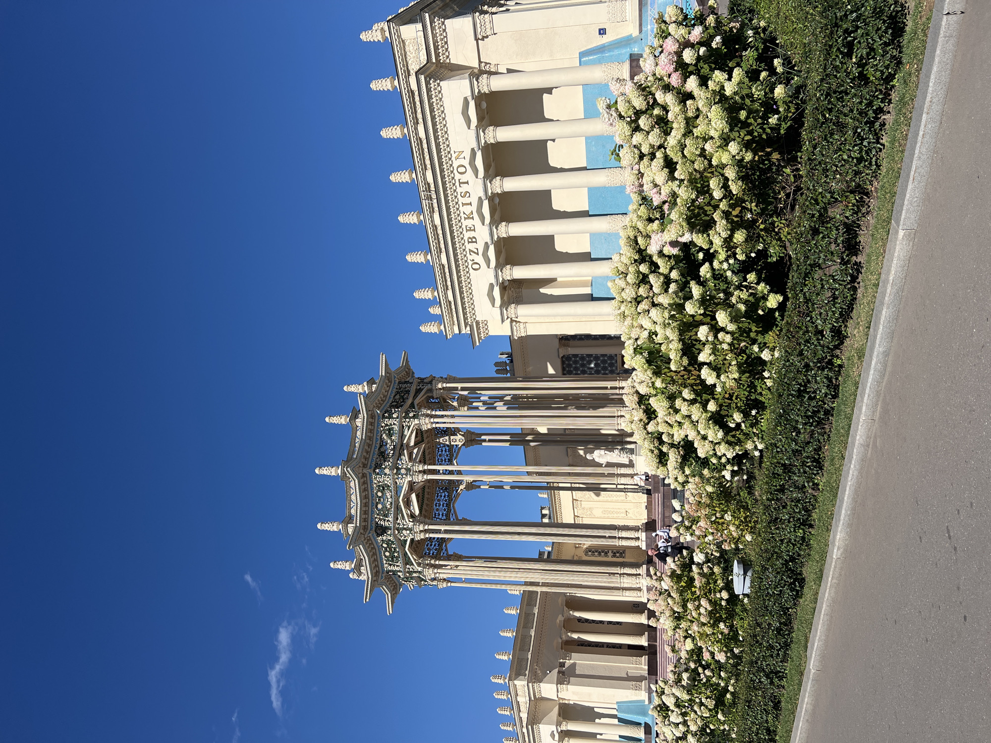 This is a photograph of the Uzbekistan Pavilion at VDNKh (Exhibition of Achievements of National Economy) in Moscow, Russia. The pavilion features a grand, cream-colored building with classical columns and a large sign spelling out "O'ZBEKISTON". In front of the main building is an ornate, multi-tiered gazebo supported by numerous columns. A dense hedge of white and pink hydrangeas runs along the front of the pavilion, with a neatly trimmed green hedge below it. A paved walkway or road is visible in the foreground, separated from the landscaped area by a concrete curb and a strip of grass. The sky is a clear, vibrant blue with a few wispy clouds. Several people are visible on the steps of the pavilion and near the gazebo. The lighting suggests it is a bright, sunny day.