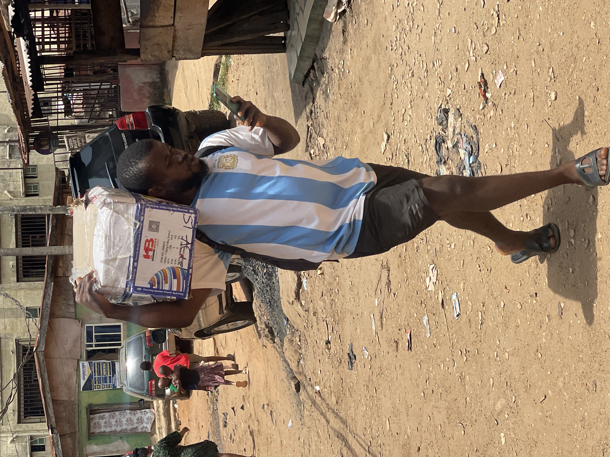 The image captures a bustling street scene in Onitsha, Nigeria, under bright, sunny daylight. The primary subject is a man walking on a dusty, unpaved road. He wears a light blue and white striped Argentina football jersey, black shorts, and dark slippers. He carries a large, rectangular package, partially wrapped in plastic, under his left arm while simultaneously looking at a smartphone held in his right hand.
In the background, a black car and a motorbike are visible. Further down the street, several people, including children and an adult in a red shirt, are gathered. The surrounding environment features multi-story buildings with barred windows and corrugated metal roofing, indicative of an urban setting. The ground is a light brown dirt road scattered with small debris.
Notable details include the clear shadow cast by the man, suggesting midday sun. Text visible on the package includes "HB HOME BEST," "ITEM NO H6012," and "DESC: 1 Gang 2 Way Switch," along with "QTY," "PCS," "GW," and "KG," suggesting it contains electrical components. The man's jersey proudly displays the "AFA" (Argentine Football Association) logo. A sign on one of the buildings reads "QUEEN BEAUTY" and "capire."