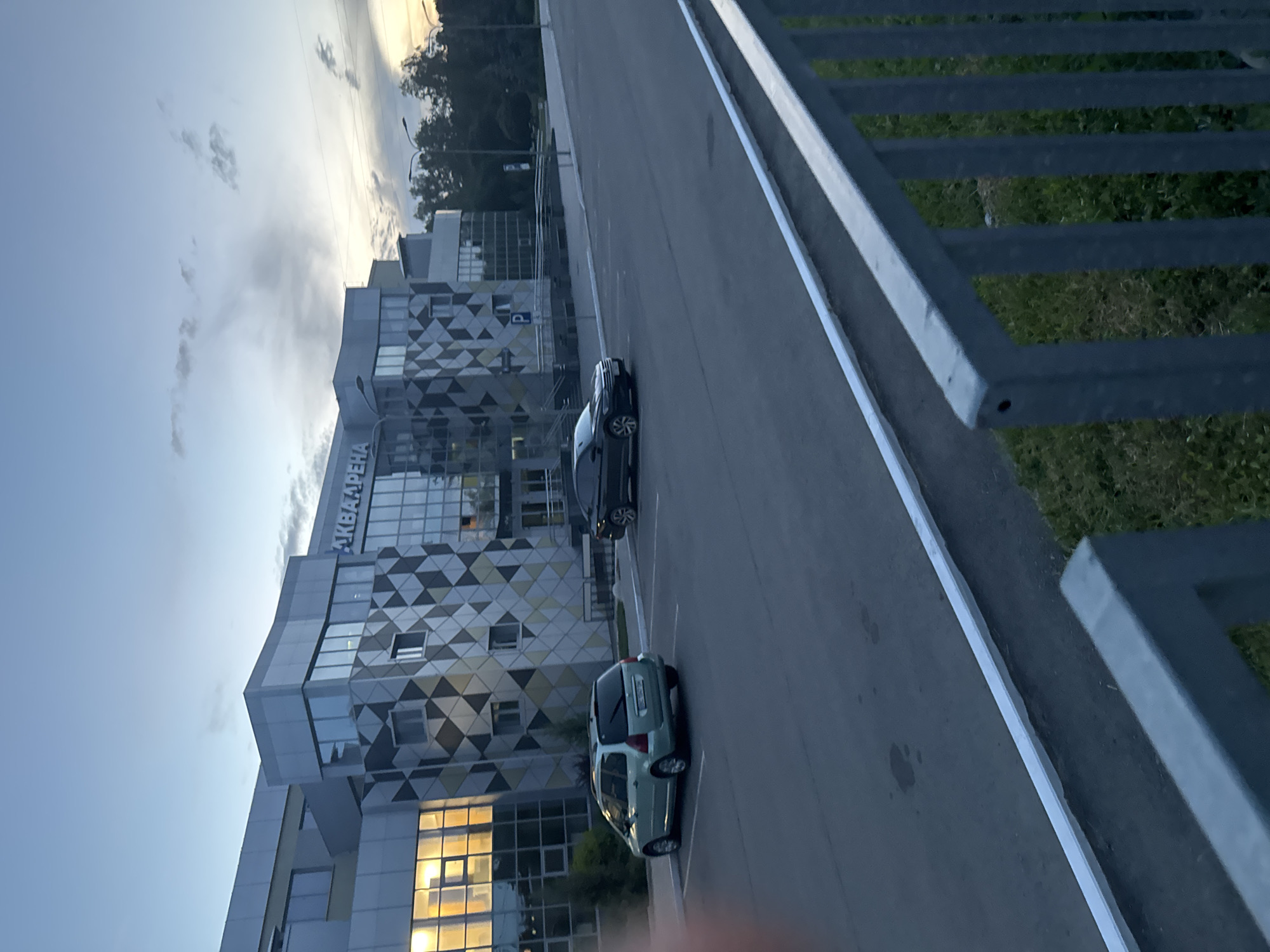 In the city of Unknown, Unknown, an exterior view captures a modern building complex with a sign reading "АКВААРЕНА" (Aquarena). The building features a facade with geometric patterns of silver, gray, and beige squares, interspersed with large glass windows. To the left, a green station wagon is parked in a designated parking space. To the right, a black sedan is parked. The scene is set in the evening, with a dusky sky overhead and streetlights visible on poles. A metal fence runs along the foreground of the image, partially obscuring a patch of green grass.