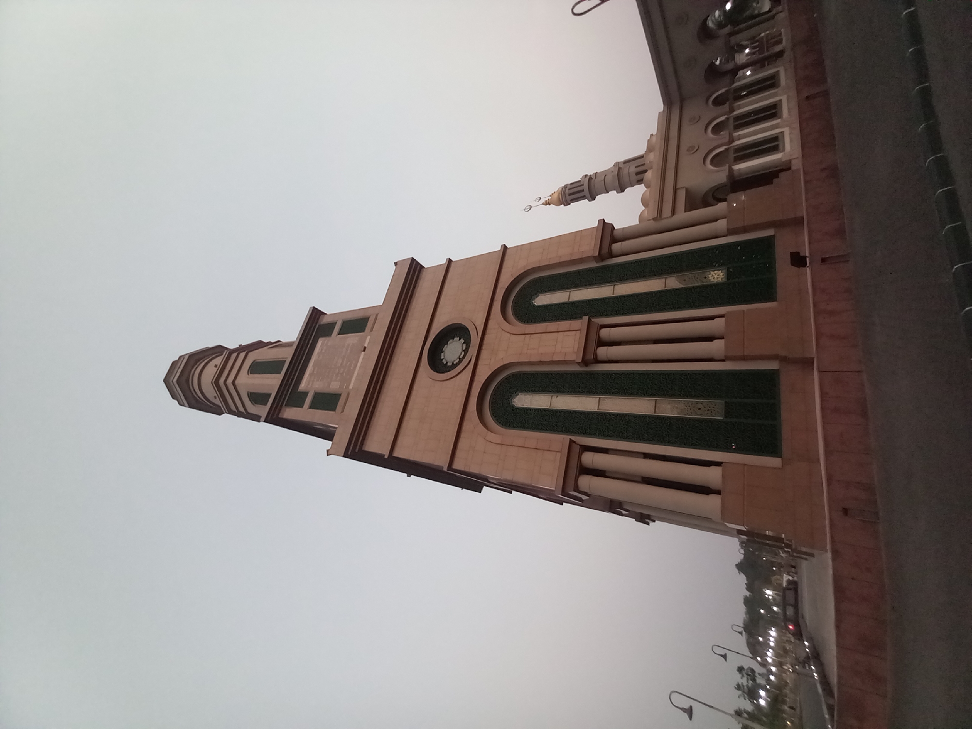 The image shows the Islamic Center Mosque in Samarinda, Indonesia. A tall, multi-tiered minaret with a golden spire dominates the center of the frame. The minaret is made of light-colored stone and features decorative green panels and arched windows. To the right of the main minaret, a smaller, adjacent minaret is visible, also topped with a golden spire. The mosque complex appears to be made of the same light-colored stone, with arched doorways and windows. In the foreground, there is a road or pathway and a low, reddish-brown wall. Streetlights are visible in the background, suggesting it is either dusk or dawn, or the area is well-lit. The sky is overcast and a pale gray. No people are clearly visible in the image.