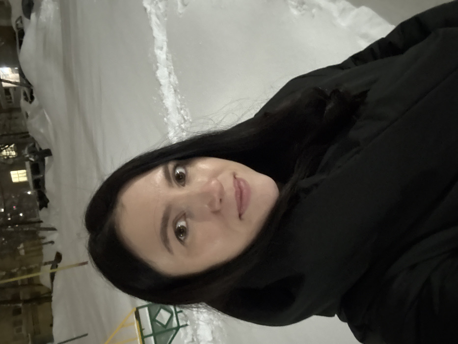 A solitary adult female is depicted in a close-up, upward-angle outdoor self-portrait, consistent with a selfie. She has long, dark hair parted centrally, brown eyes, and a fair complexion, displaying a neutral expression with a slight smile. She is wearing a black hooded winter coat with a high collar, obscuring her body below the mid-chest.
The scene is an urban residential area at night, under artificial illumination, covered in deep, undisturbed snow. To the subject's left, a portion of a yellow and green metal playground structure is visible, partially buried in snow. In the background, several parked vehicles are heavily covered with snow. Beyond the vehicles, multi-story residential buildings are visible, with at least one illuminated window emitting a warm, yellow light. Bare trees or bushes are also present in the snow-covered landscape. Footprints are discernible in the snow to the right foreground. The environment is consistent with a winter night in a residential district of Kazan, Russia, though no specific landmarks are visible to pinpoint an exact address or neighborhood within the city.