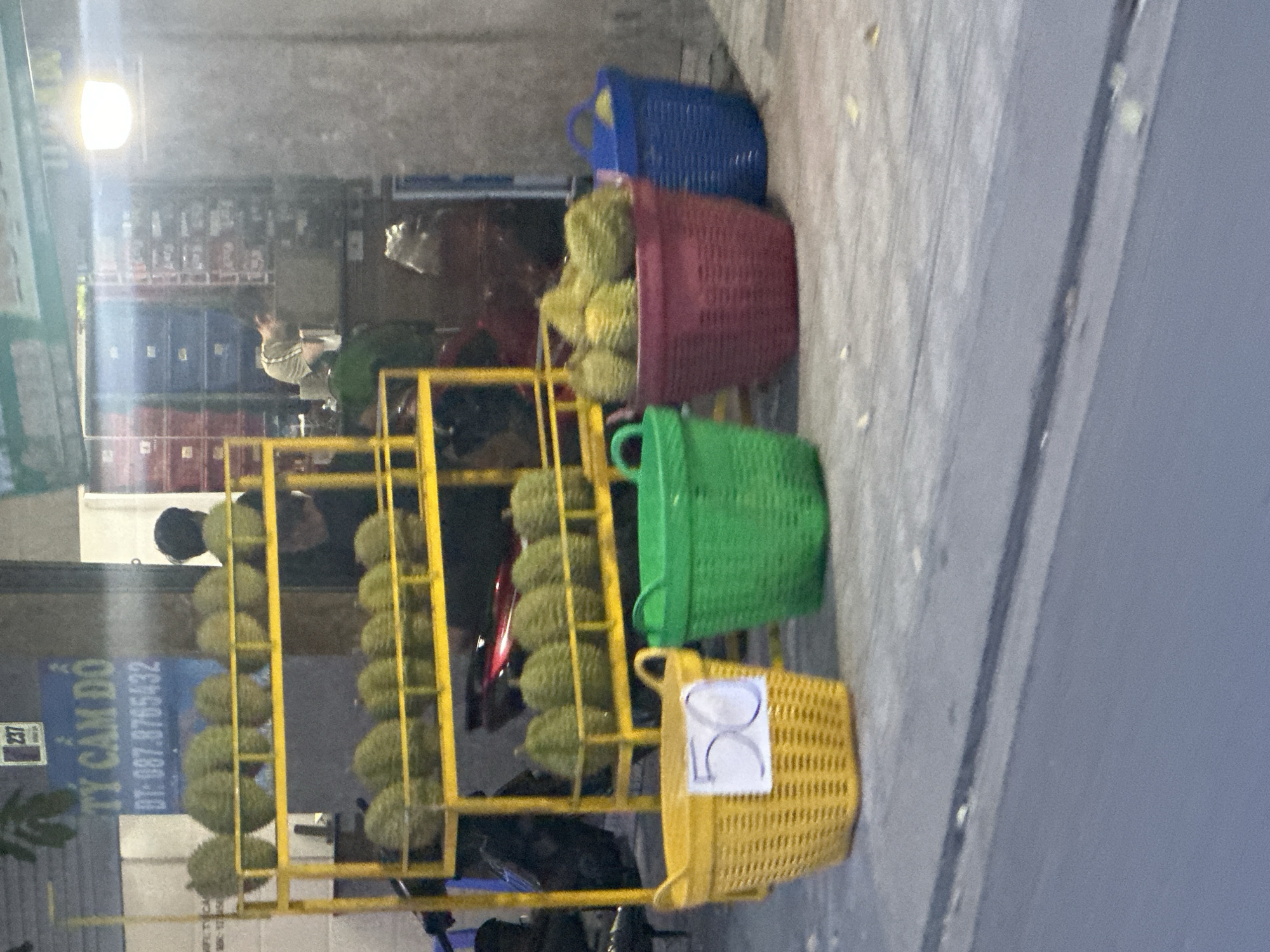 The image shows a street scene in Bac Nha Trang Ward, Vietnam. In the foreground, a yellow metal rack displays two columns of durians. The rack is situated beside a motorbike. Four baskets are placed on top of the rack, stacked from top to bottom in the following colors: blue, red, green, and yellow. The yellow basket has a white sign attached, with a price written on it. A shop sign, written in Vietnamese, and with a phone number on it, is visible in the background, slightly blurred. A person is standing near the durians. In the far background, the interior of the shop is visible. The image is angled. The street surface is gray concrete.