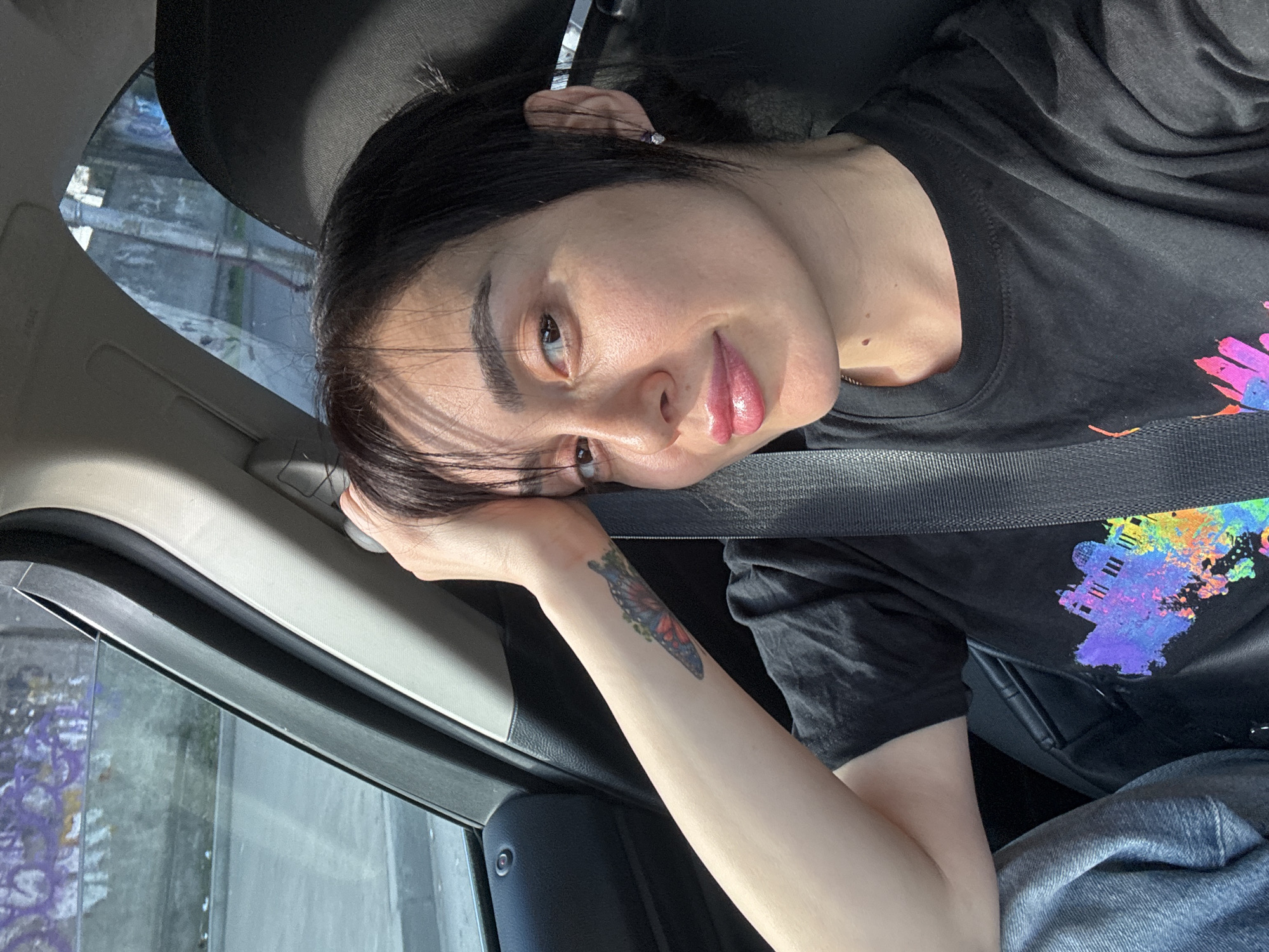 A woman is in a car, likely a passenger, wearing a black T-shirt with a multicolored design and a seatbelt. She has dark hair and fair skin, with a butterfly tattoo on her forearm. She is leaning her head against the car frame with her hand. The background shows an urban environment with buildings, possibly in Tbilisi, Georgia. The scene is captured with a bright, natural lighting condition. The car window shows a blurred outdoor setting.