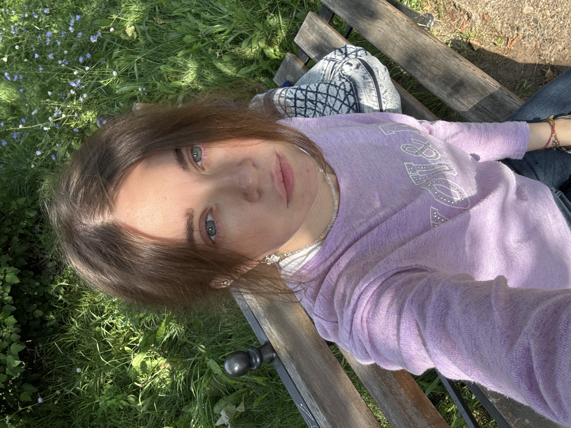 A young woman with brown hair and blue eyes is sitting on a wooden park bench. She is wearing a light purple long-sleeved shirt adorned with rhinestones spelling out "Got&lo", and blue jeans. She is looking directly at the camera, with her head slightly tilted. A patterned backpack is visible behind her on the bench. The background consists of lush green grass, foliage, and small blue wildflowers, suggesting an outdoor, natural setting. The lighting indicates it is daytime, with sunlight casting shadows on the bench and illuminating the woman's face. The overall scene appears to be a casual moment captured in a park.