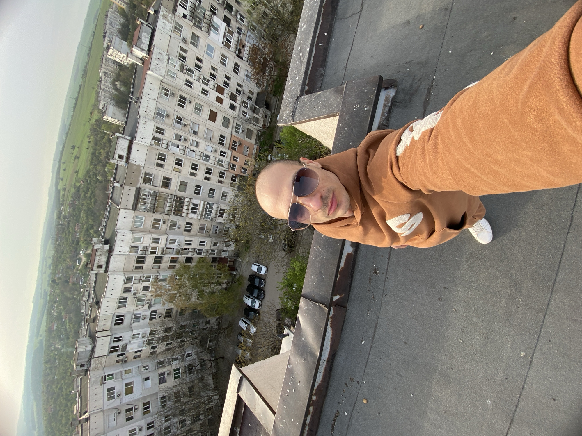 A man is standing on the edge of a high-rise building, taking a selfie. He is wearing a brown hoodie and sunglasses. The background shows dense multi-story apartment buildings and a green landscape, suggesting a location in Pleven, Bulgaria. Cars are parked in the courtyard of the building below. The surface beneath the man appears to be a flat, grey rooftop.