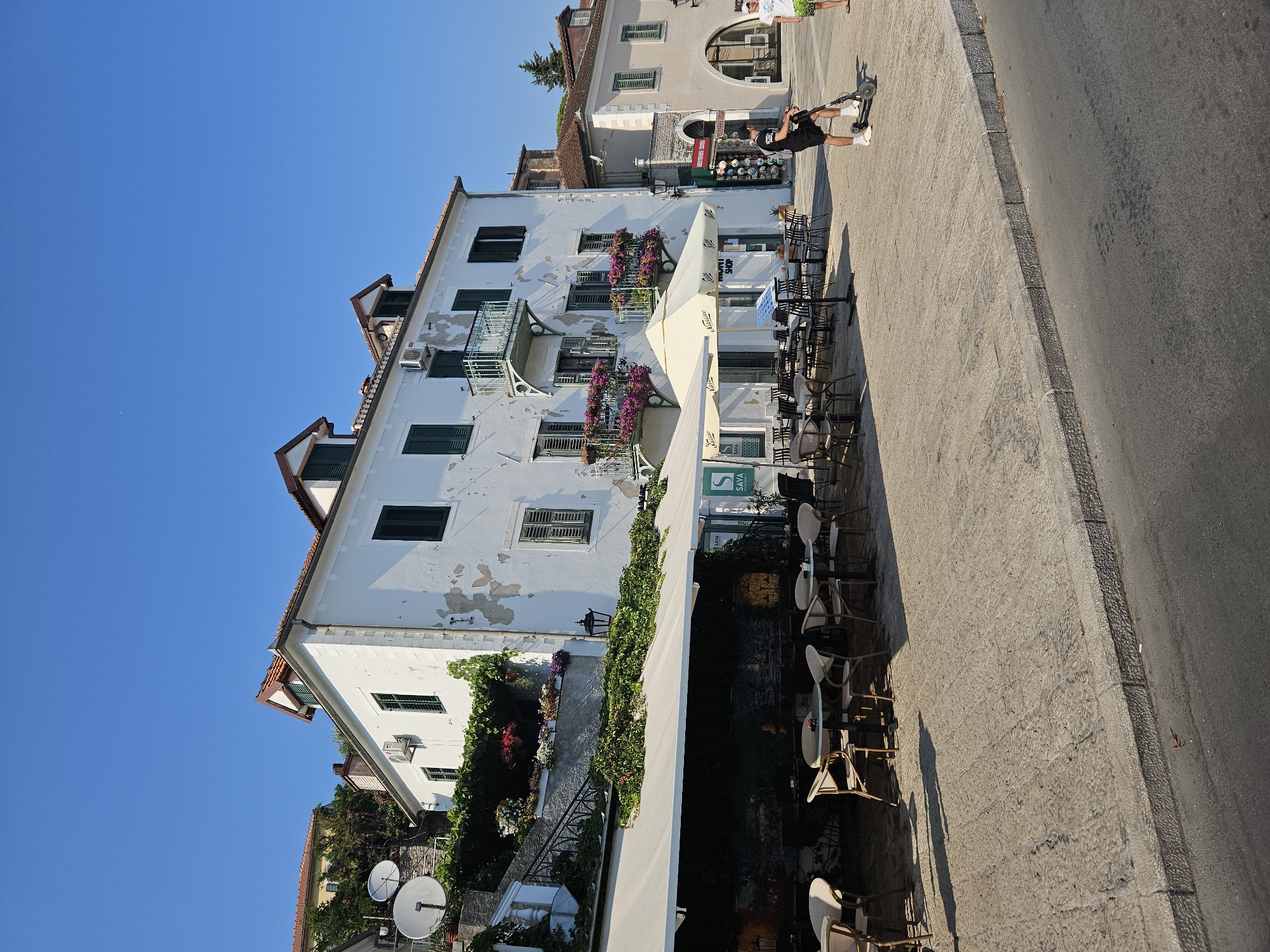 The image depicts a sun-drenched, outdoor scene in Herceg Novi, Montenegro. A white building with peeling paint and green shutters dominates the center of the frame. The building has balconies adorned with vibrant pink flowers and overlooks a paved square in front of an outdoor cafe. Several tables and chairs are set up under a large white awning, with some patrons seated and others absent.
To the left of the main building, a steep, stone staircase winds upwards, bordered by lush green ivy and potted plants. Two white satellite dishes are mounted on the hillside behind the staircase, suggesting a residential or communication hub.
In the foreground, a man on an electric scooter rides across the paved square, wearing a black t-shirt and shorts. Another person in a white t-shirt and green shorts stands to the right, facing away from the camera. The square is paved with large, irregular stones, and a grey concrete curb separates it from an asphalt road. The sky is a clear, bright blue.
In the background, another building with an arched entryway is visible, featuring an assortment of goods displayed in its windows, possibly indicating a shop. The overall impression is one of a lively Mediterranean town on a clear, warm day.