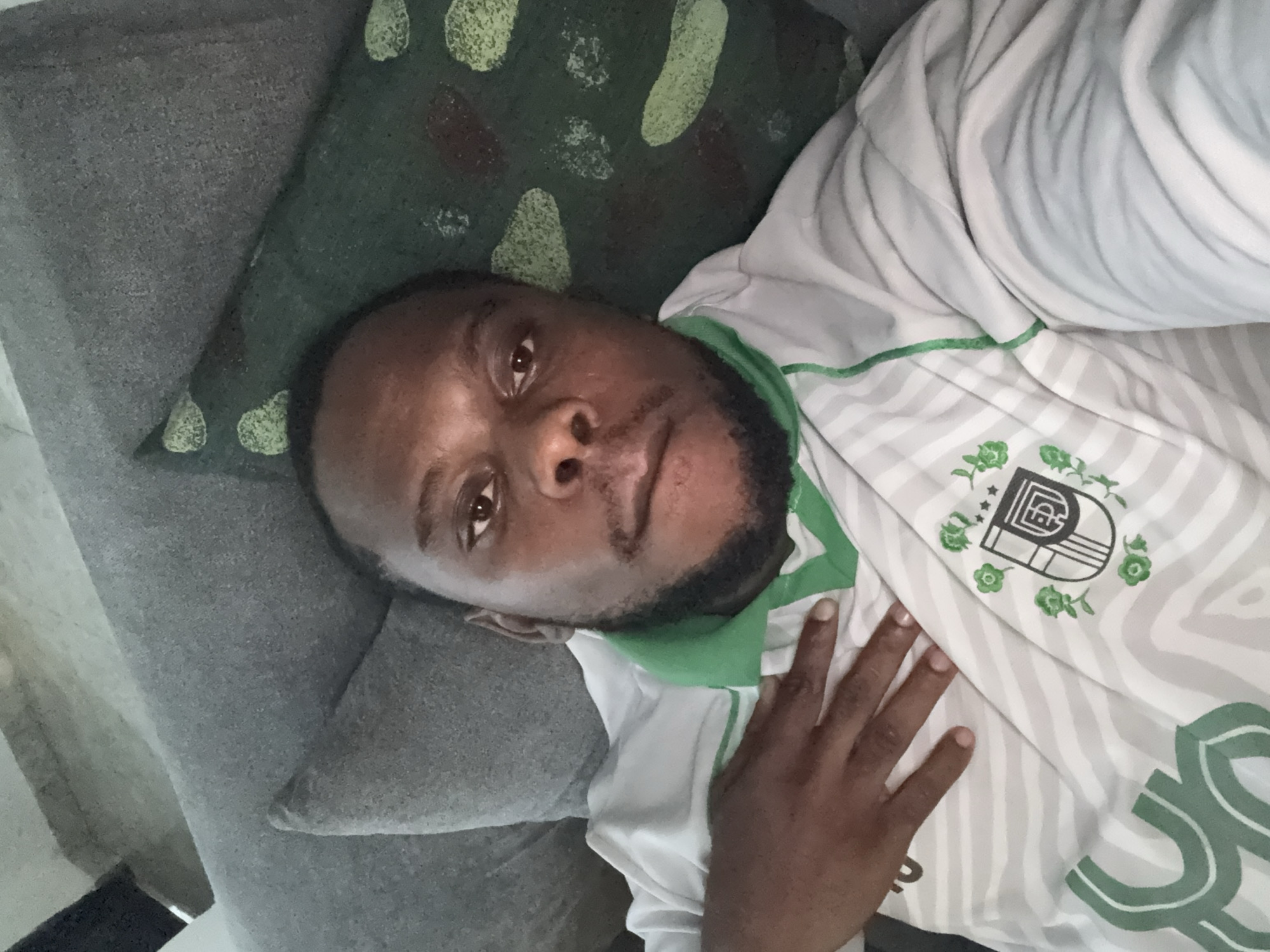 A man lies on a grey couch, his head resting on a green patterned pillow. He is wearing a white shirt with grey stripes and a green collar. A crest with green floral accents is visible on the shirt. The man is dark-skinned with a beard, and his hand rests on his chest. The location is not discernible from the image. No specific information indicating Iddo Tudunwada, Nigeria is available.