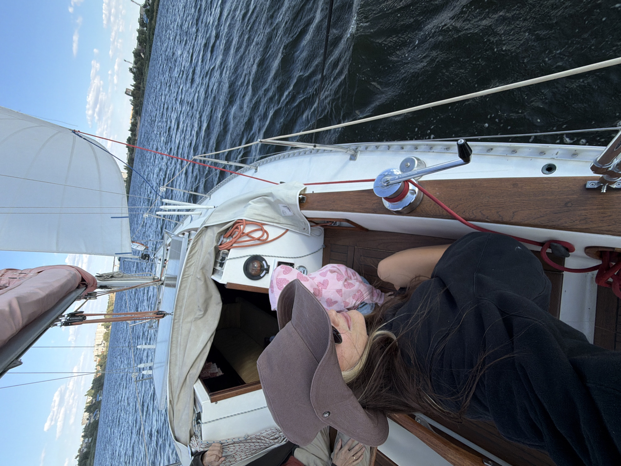 The image shows a person wearing a wide-brimmed hat and sunglasses lying on a sailboat. The person is wearing a black hoodie. Behind the person, a small child in a pink hoodie with hearts is visible. The sailboat is on a body of water, likely the Volga River, with the city of Kamyshin, Russia, visible in the background. The sky is blue with some clouds. The scene appears to be set during the daytime.