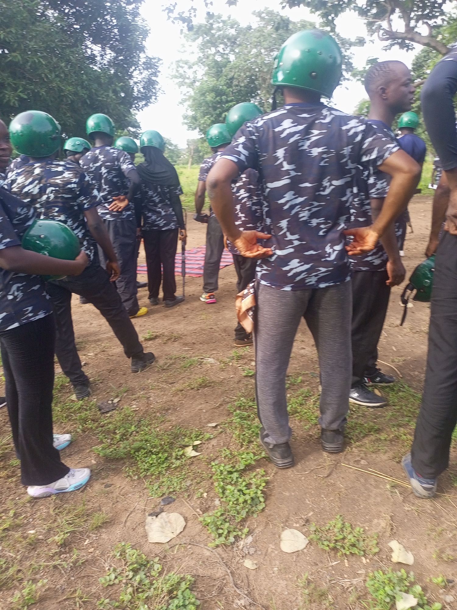In Takum, Nigeria, a group of individuals, likely police officers based on their attire and green helmets, are gathered outdoors on a dirt and grassy area. The people are wearing camouflage patterned shirts and dark trousers. Several individuals are wearing green helmets. Some are holding green helmets, while others have them on their heads. One person is holding a long, dark object that could be a rifle. The environment appears to be natural with trees visible in the background, suggesting an open, possibly rural or training, area. The lighting suggests it is daytime.