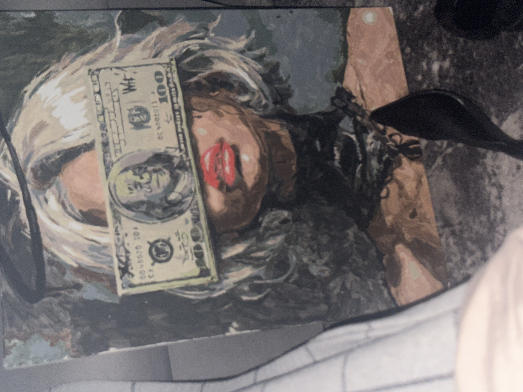 The image shows a painting of a woman with blonde hair and red lips. A one hundred dollar bill is placed across her face. The painting rests on a surface that appears to be a bed or couch, with partial views of a white and possibly gray patterned fabric. The location in Высокий, Russia, is not discernible from the image. There is also a high heel in the image.