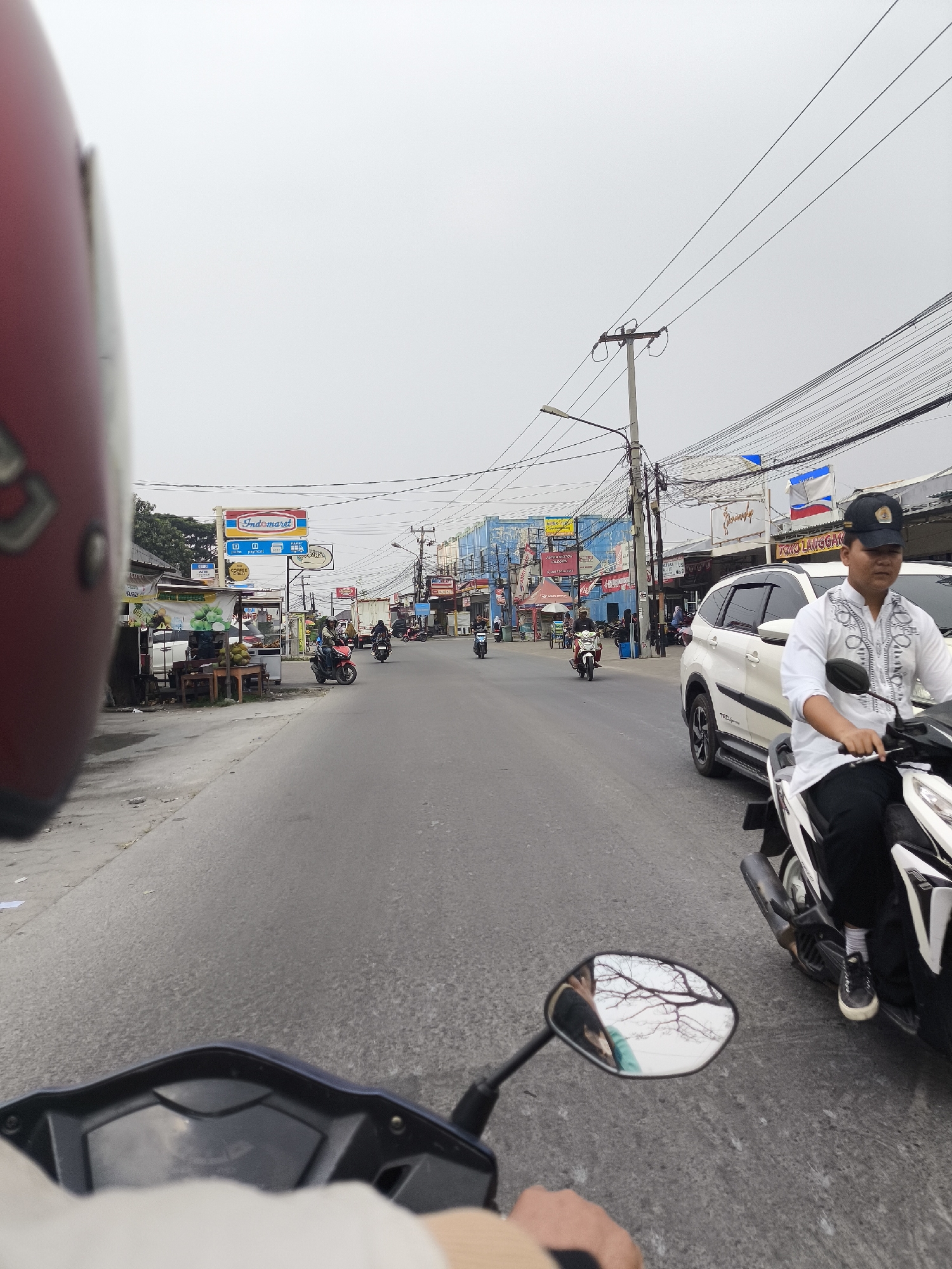 This is a first-person perspective image, taken from the viewpoint of someone riding a motorcycle in Cilegon, Indonesia. The foreground shows the handlebar and part of the dashboard of a dark blue scooter, with a person's hand visible gripping the handlebar. The rider is wearing a light-colored long-sleeved shirt. To the left, a portion of a red helmet is visible, indicating the rider is wearing one.
The motorcycle is on an asphalt road, which appears to be a street in a commercial area. On the right side of the road, a white SUV is parked, and a man wearing a white shirt with black trim and a black baseball cap is riding a white scooter. He is wearing black pants and white sneakers. The scooter he is riding is mostly white with black accents.
The left side of the road features several small businesses and stalls. A prominent sign for "Indomaret," a popular convenience store chain in Indonesia, is visible, along with other signs for shops and services. There are trees and some foliage visible behind the buildings. Several other motorcycles and a few pedestrians are present on the road and sidewalks.
In the background, a utility pole with numerous electrical wires stretches across the sky, a common sight in Indonesian urban environments. The sky is overcast and light gray, suggesting a cloudy day. The overall scene depicts a typical bustling street in a city, with moderate traffic and commercial activity.