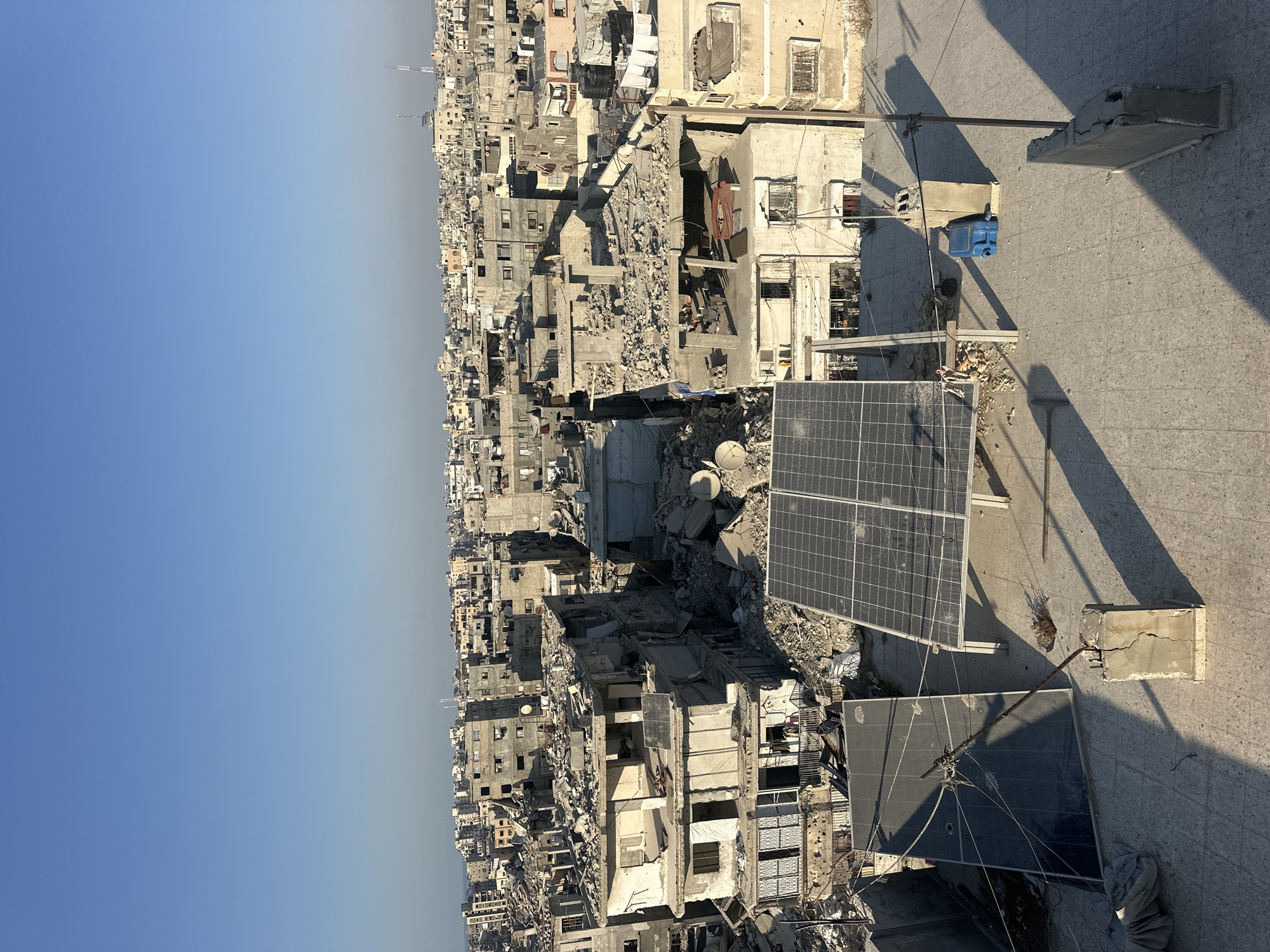 This image depicts a widespread scene of destruction in the city of Gaza, Palestinian Territories. The foreground is dominated by a rooftop with two solar panels, one standing upright on a metal frame and another lying flat, partially covered by debris. A broken concrete post, cracked and weathered, supports a rusted metal pole connected to wires. Shadows cast by the solar panels and poles stretch across the tiled rooftop.
In the midground and background, a densely populated urban area is visible, characterized by extensive damage to buildings. Structures appear to be partially or completely collapsed, with exposed rebar, rubble, and missing walls. Many buildings show signs of structural failure, with floors caved in and roofs destroyed. Satellite dishes are visible on some of the damaged buildings. The overall impression is one of severe destruction and devastation. The sky is a clear, bright blue, contrasting sharply with the grim reality of the urban landscape below.