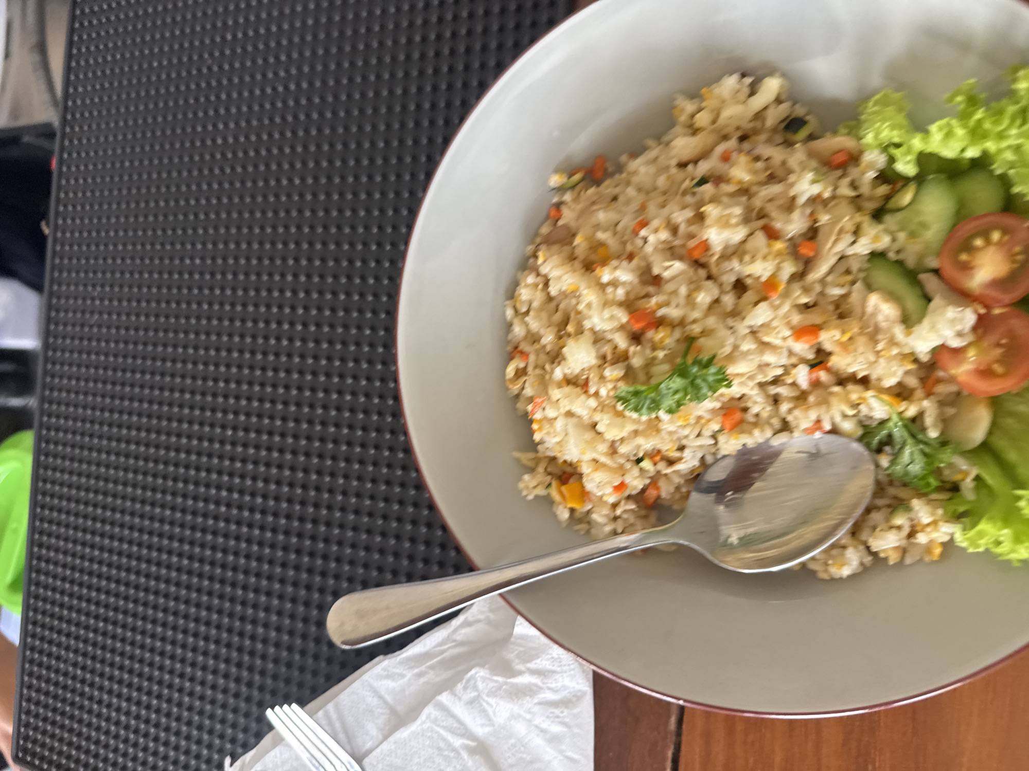A high-angle shot captures a plate of fried rice with a side salad, placed on a table in Denpasar, Indonesia. The fried rice, featuring visible carrots and other vegetables, dominates the upper right of the frame. A silver spoon rests on the rice, near the center of the plate. The side salad, consisting of lettuce, sliced cucumbers, and halved cherry tomatoes, occupies the right edge of the plate. The plate itself has a gray interior and a thin red rim. The table is wooden and of a light brown color. A black, textured mat is to the left of the plate. A crumpled white napkin and a portion of a fork are visible in the lower left corner. No people are visible in the image.