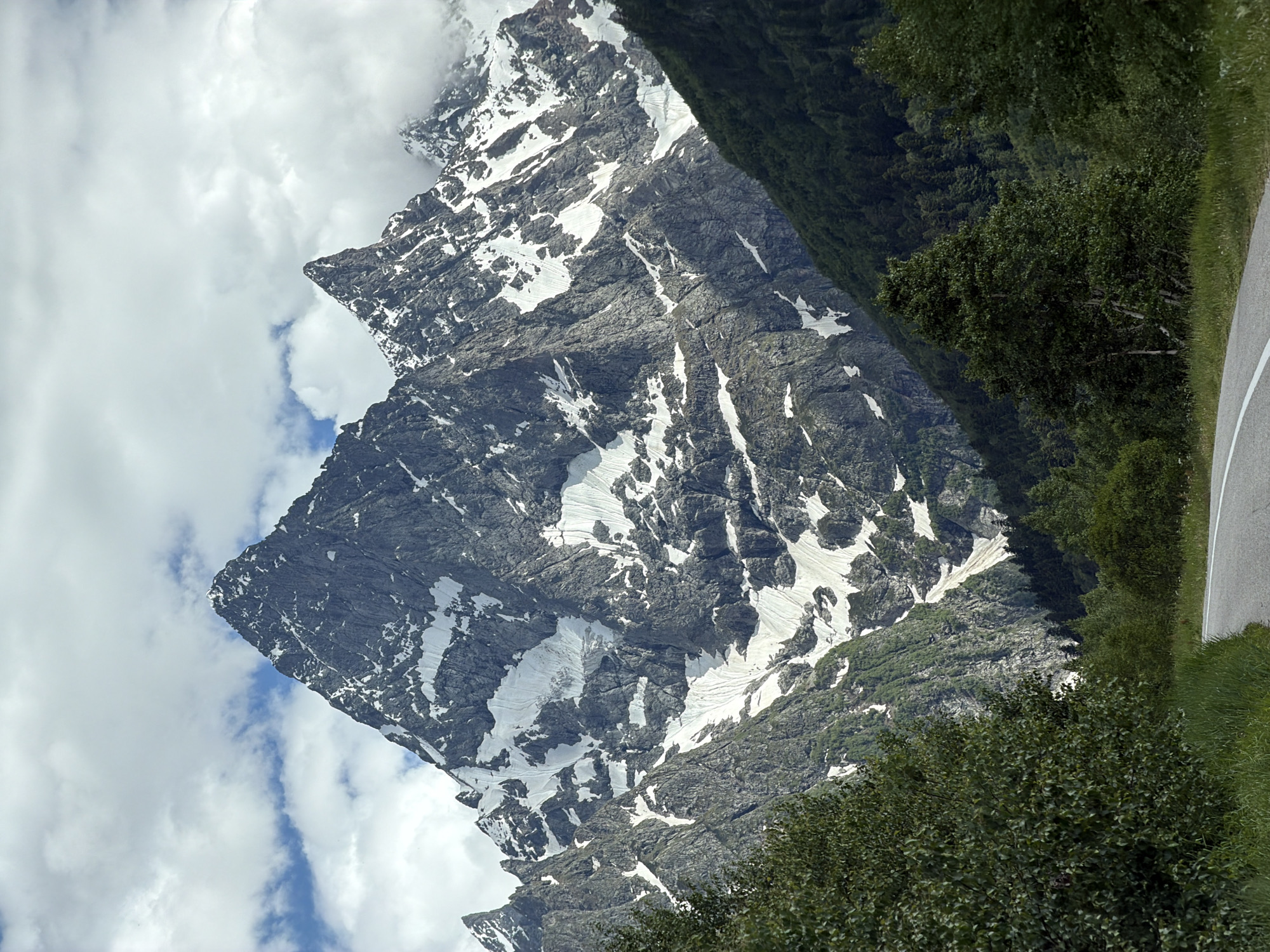 A wide-angle, eye-level, outdoor shot captures a majestic mountain range under a cloudy sky. The dominant feature is a massive, jagged mountain peak, partially covered in snow, with steep rocky slopes. To its right, a smaller, sharp peak pierces the clouds. The lower slopes of the mountains are covered in lush green trees, forming a dense forest. In the foreground, a paved road with white lane markings curves from the bottom left towards the right, bordered by grassy verges. The scene is tranquil and untouched, suggesting a remote, natural setting. The location is identified as Верхне-Тебердинское сельское поселение, Russia. There are no people or animals visible in the image.