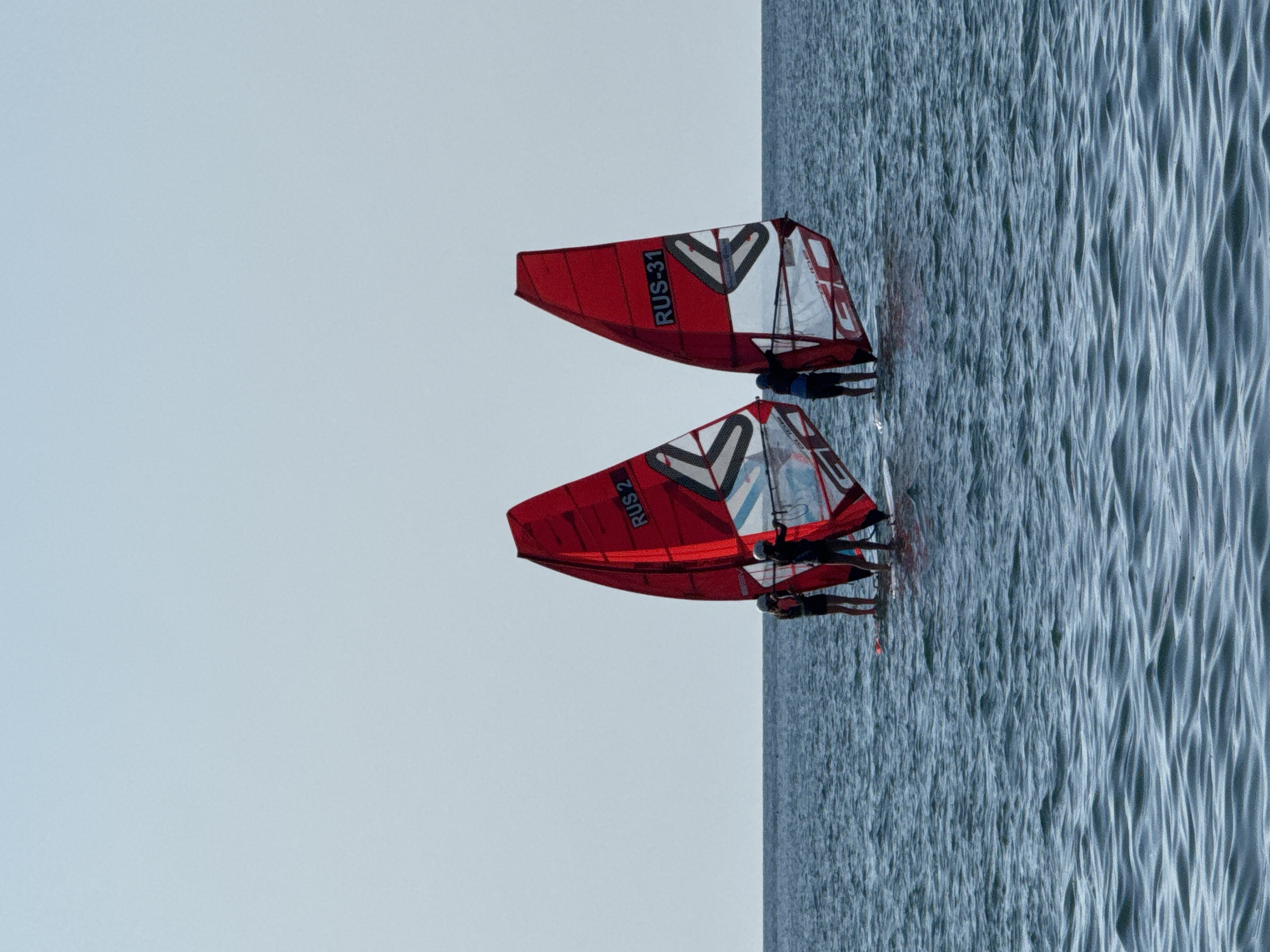 The image depicts a serene marine scene with two individuals windsurfing on a calm body of water. The sky is a clear, light blue, and the horizon is sharply defined by the meeting of the sea and sky. The water is a deep blue, with subtle ripples suggesting a gentle breeze.
Dominating the foreground are two red windsurfing sails, emblazoned with white and black graphics, and carrying the designations "RUS 2" and "RUS-31" respectively. Each sail is attached to a windsurf board, on which a person is standing. The individuals are seen from the waist down, with their bodies silhouetted against the sails. They appear to be engaged in the activity of windsurfing, with one person on the left and another on the right, positioned slightly further back. The overall impression is one of a peaceful, recreational outing on the water.
The exact location within Sevastopol, Ukraine, is not discernible from the image itself. However, given the context of windsurfing, the scene is likely a coastal area or a bay within or near Sevastopol, known for its maritime activities.