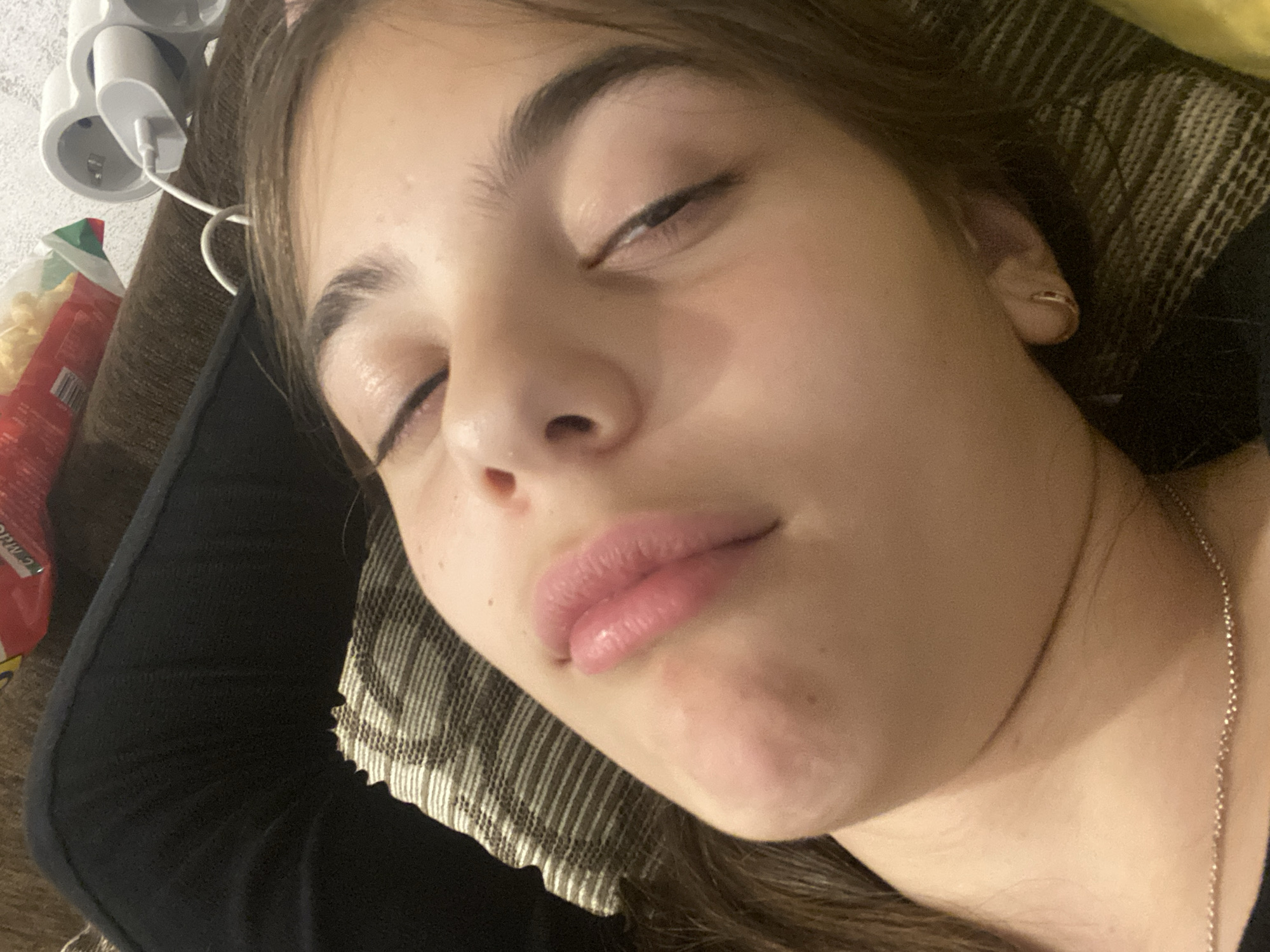 A young woman is lying down, likely resting or sleeping. She has fair skin, dark hair, and slightly parted lips. She appears to be lying on a patterned cushion or pillow, and is wearing a thin necklace. Near her head is a multi-socket power adapter and a bag of chips or snacks. The image does not contain information to confirm the location is Dragovishtitsa, Bulgaria, or any specific details about a city.