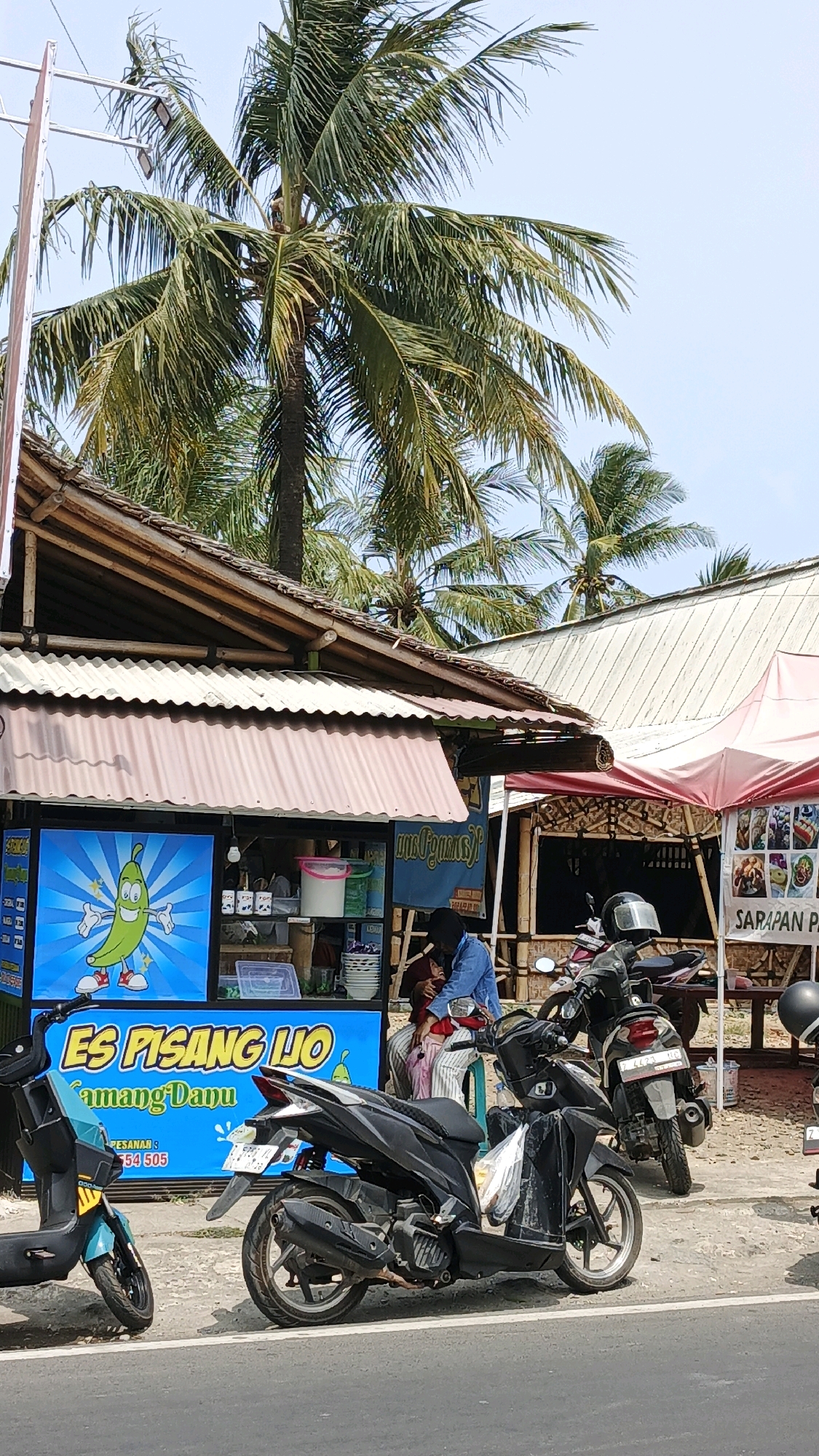 A roadside food stall in Desa Wonoharjo, Indonesia, features a bright blue sign with "ES PISANG LJO" and a cartoon banana. Behind the counter, a person wearing a headscarf and blue jacket sits with another person. Several motorcycles are parked in front of the stall, and palm trees tower overhead. A canopy extends over a portion of the stall, and a sign with images of food is visible on the right.